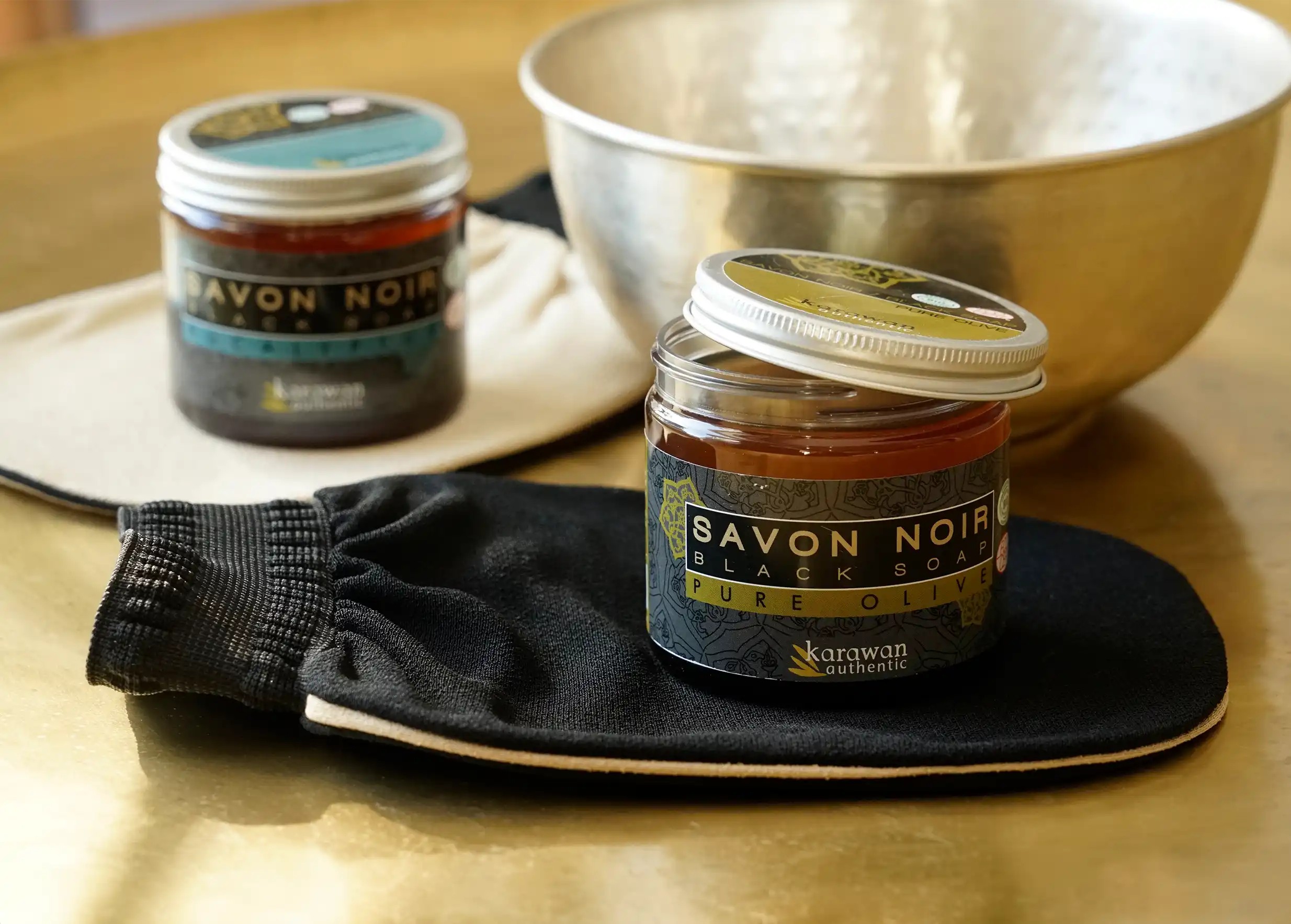 Two jars of black soap placed on kessa gloves, with a brass bowl in the background