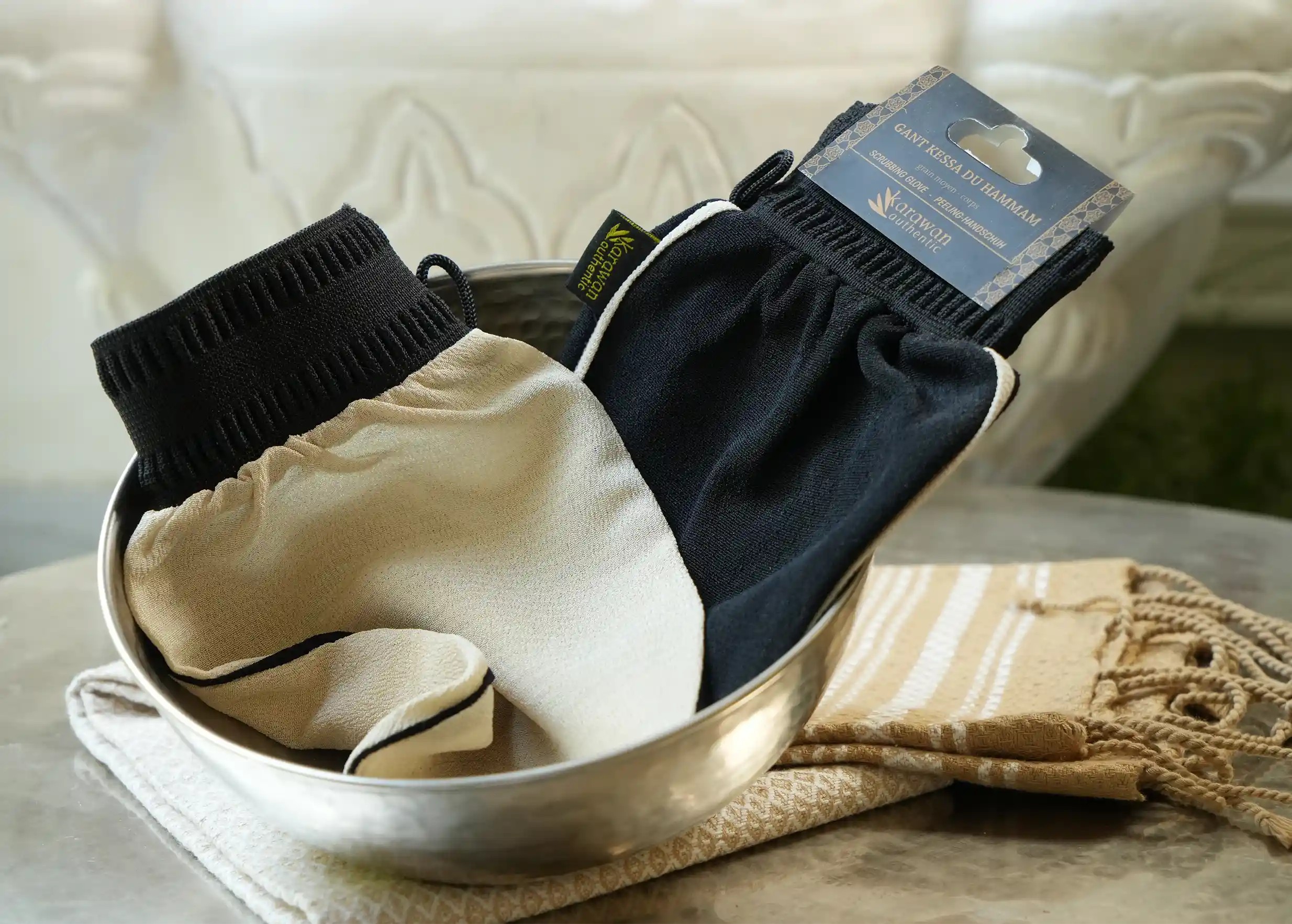 Two hammam kessa gloves, one beige and one black, set in a brass bowl on a hand-woven fouta