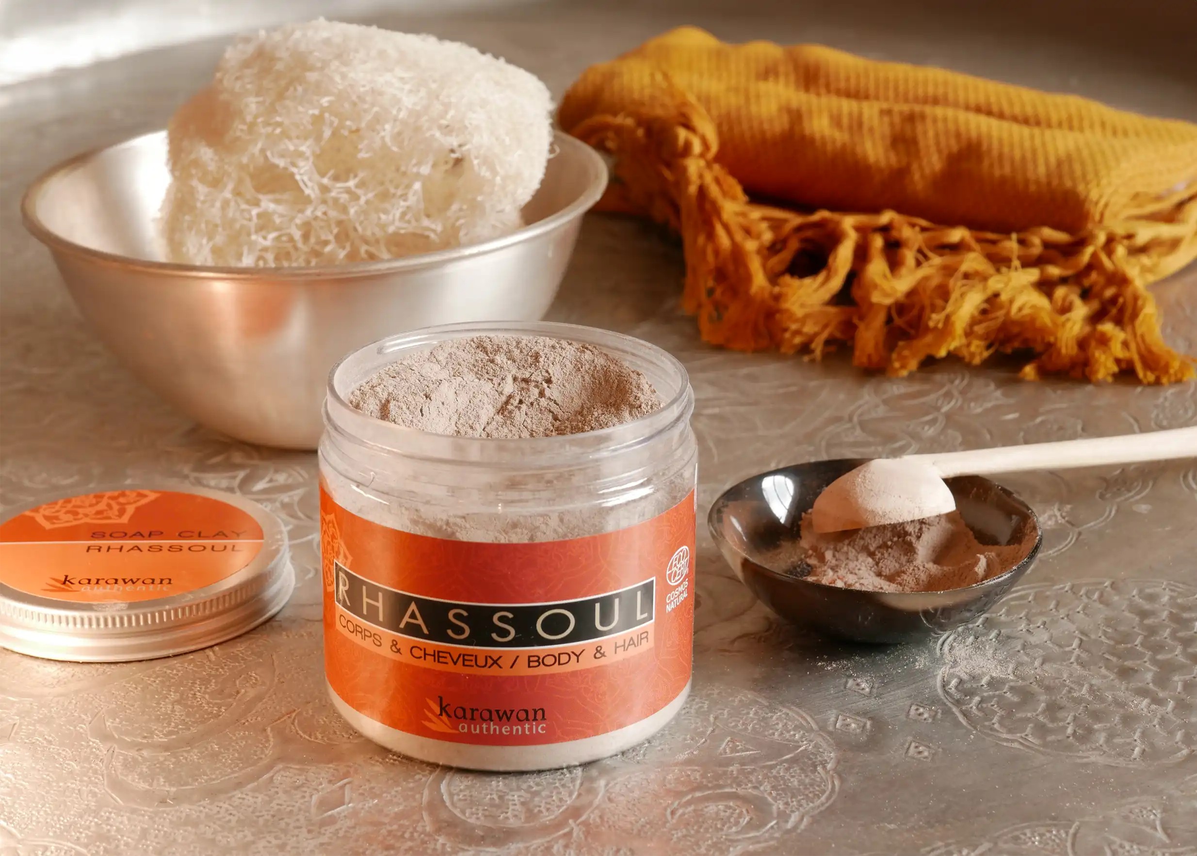 Open jar of rhassoul, Moroccan clay in a dish, vegetable sponge and yellow cotton fouta