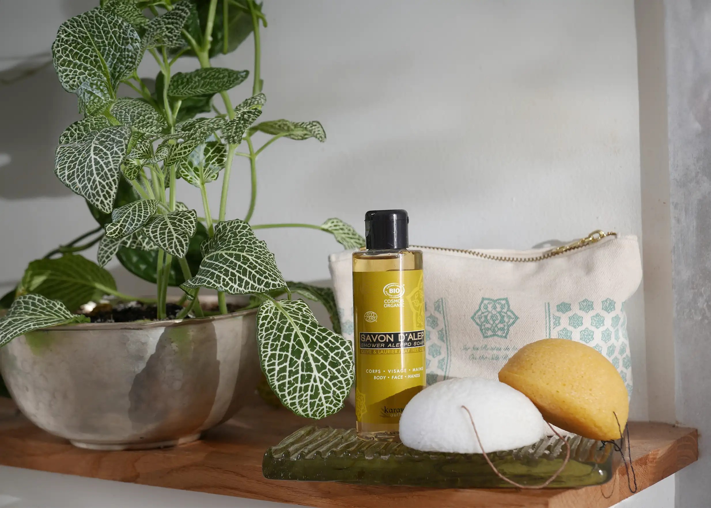 Éponge de konjac bio naturelle, entourée d'autres accessoires de bain, trousse, mini flacon de savon liquide, et des plantes