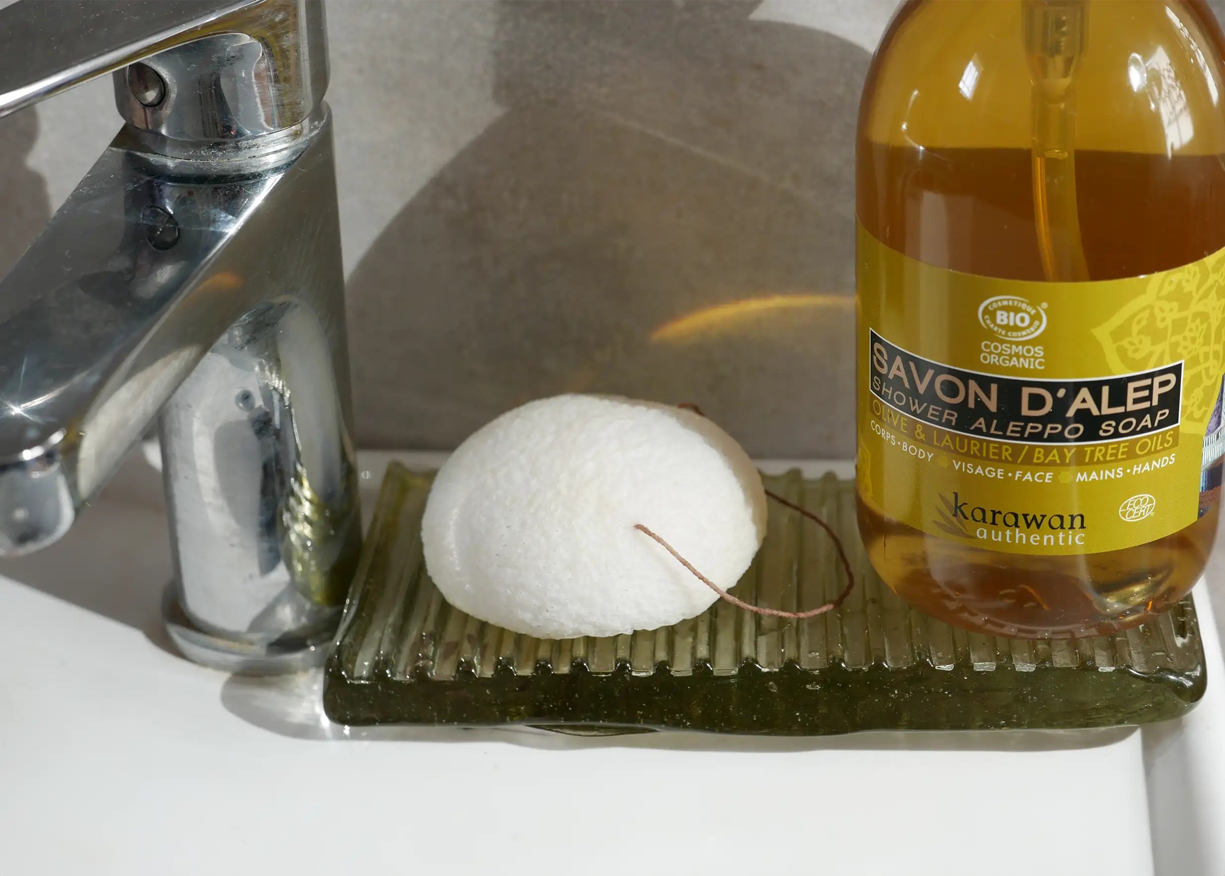 Éponge de konjac bio naturelle sur un porte savon en verre, à côté d'un savon d'Alep