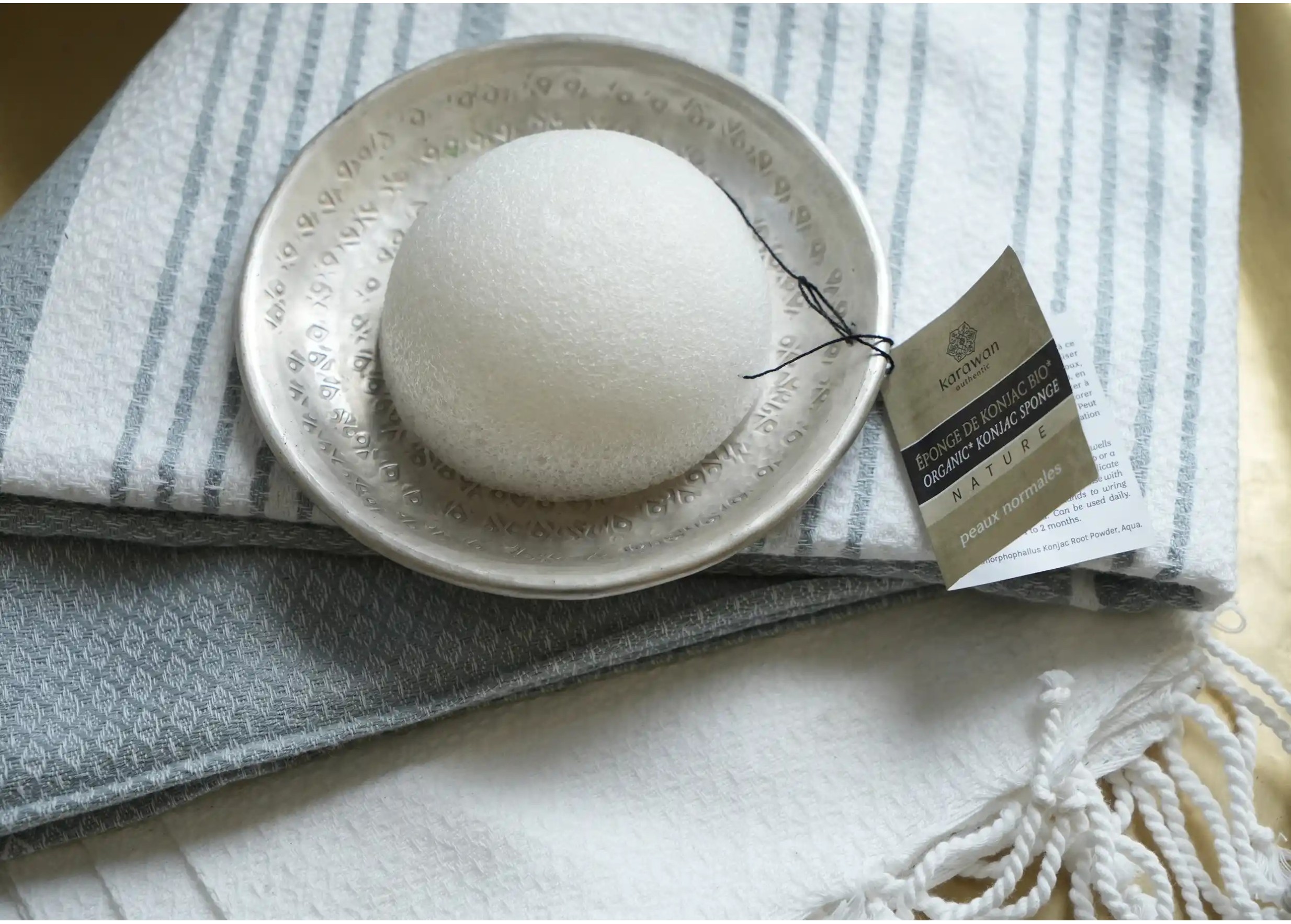 Éponge végétale de konjac naturelle au centre d'une coupelle, posée sur une fouta en coton bio