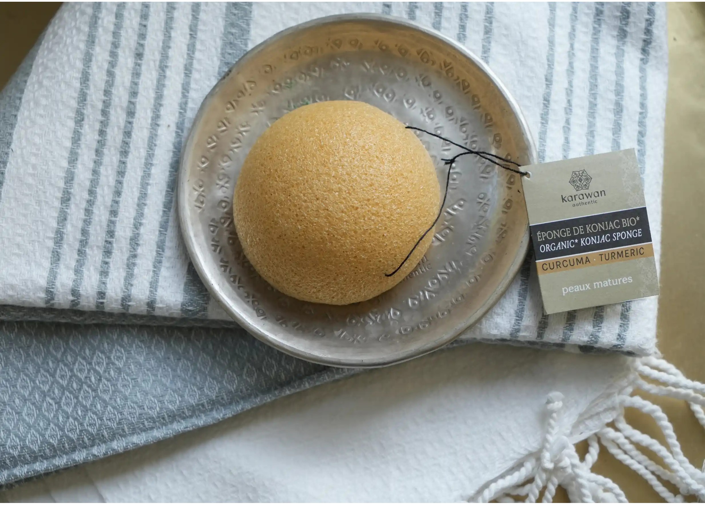 Éponge de konjac avec cordelette et étiquette carton affichant le logo Karawan posée sur une coupelle