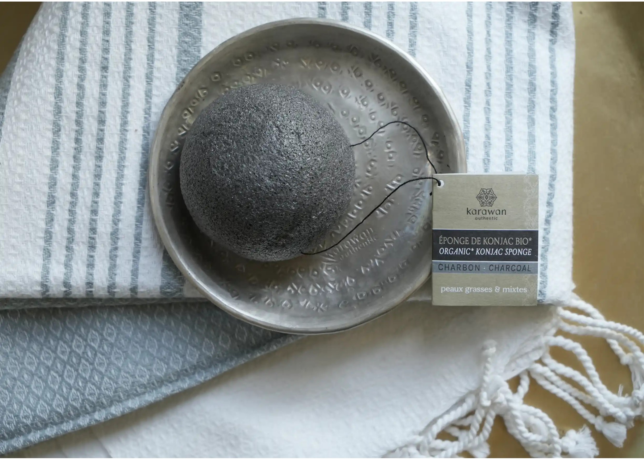 Éponge de konjac avec cordelette et étiquette carton affichant le logo Karawan posée sur une coupelle