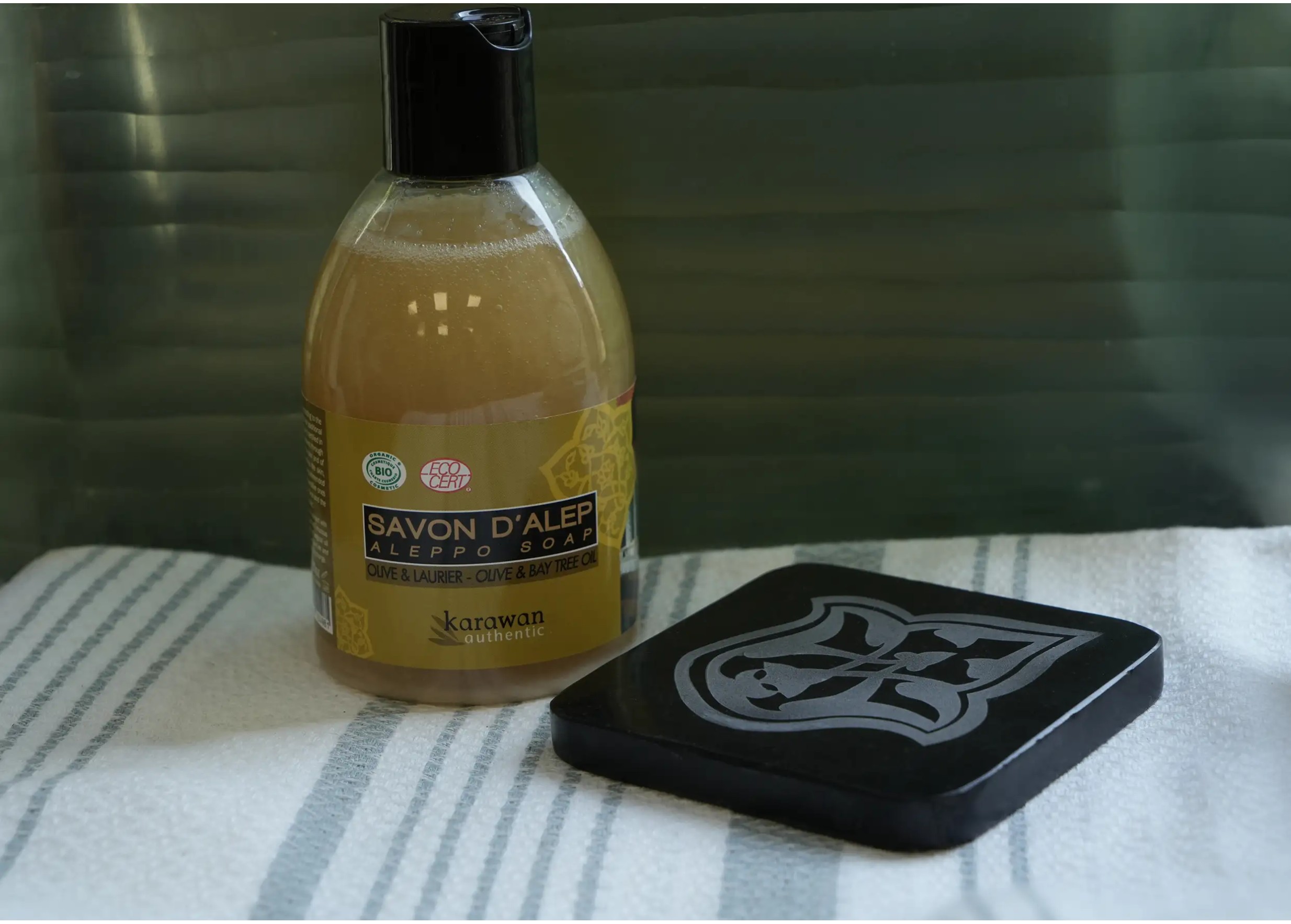 Black marble soap dish set on a fouta, next to a bottle of liquid Aleppo soap with noble olive and laurel oils.