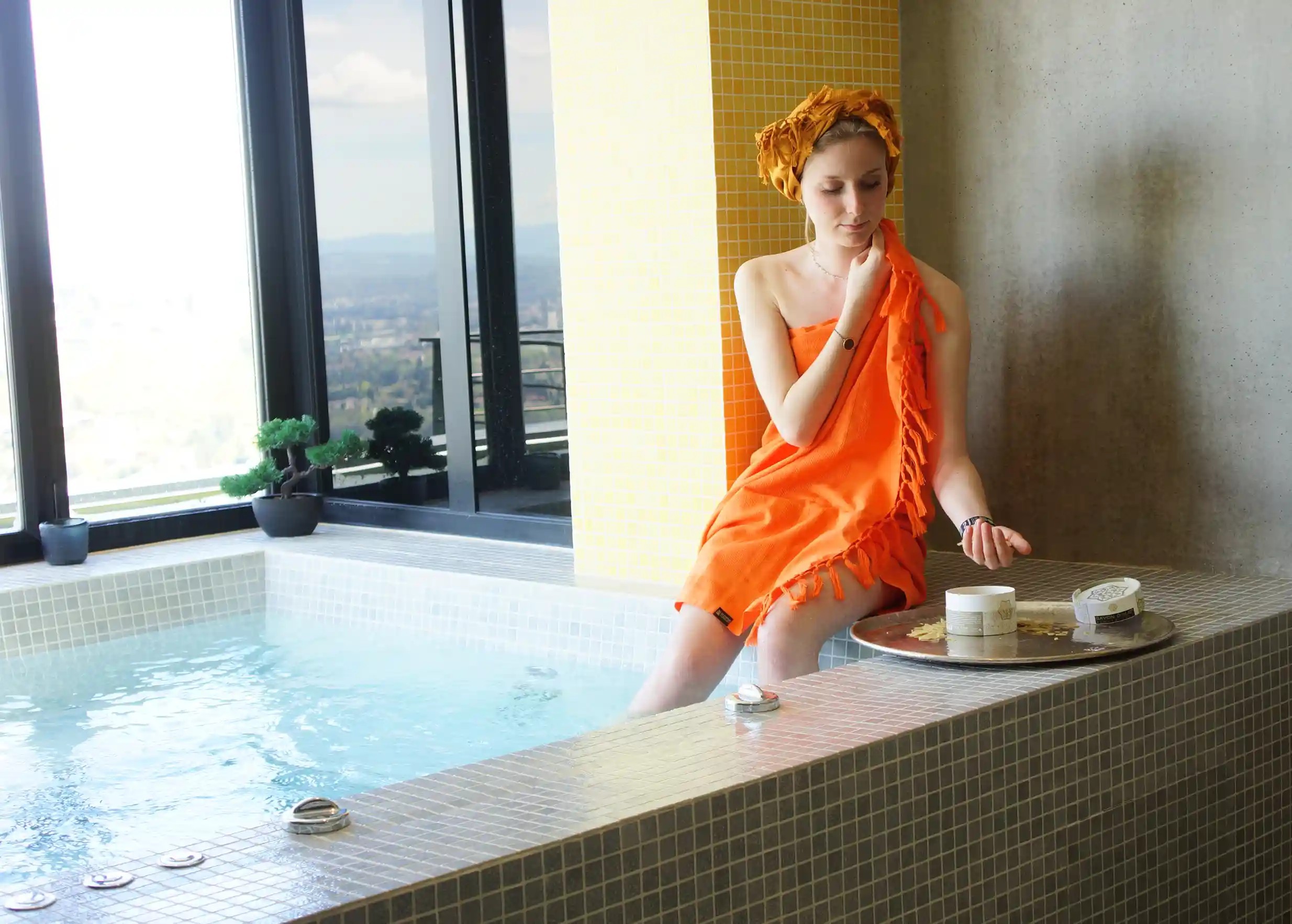 Femme assise près d'un jacuzzi, enveloppée dans une fouta en coton bio couleur curcuma, observant un plateau de produits de soin