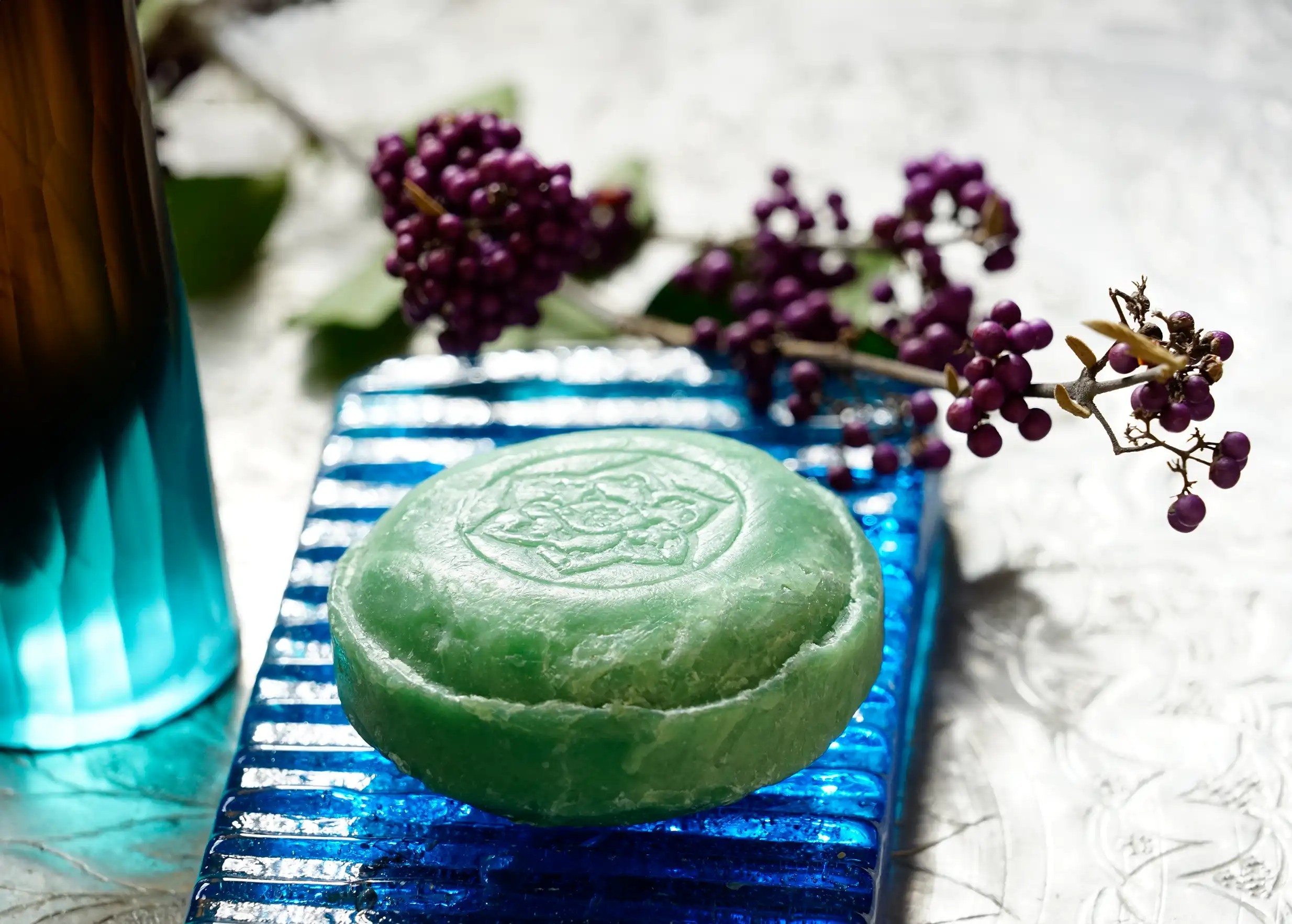 Savon ayurvédique vert au vétiver, posé sur un porte savon en verre bleu avec des branches de baies violettes en arrière-plan