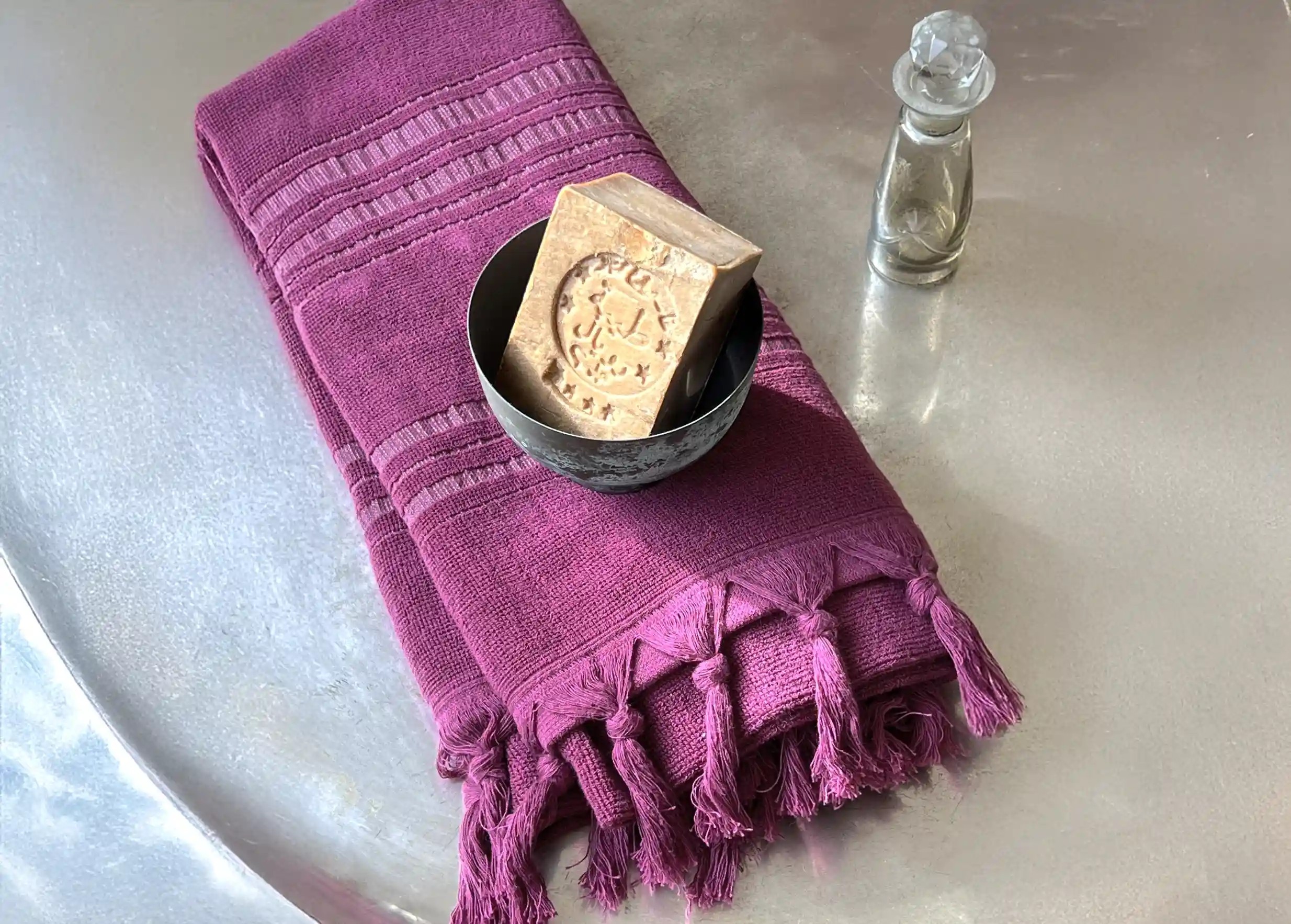 Amethyst-coloured dolce fouta in organic cotton, accompanied by Aleppo soap in a bowl, both set on a brass tray.