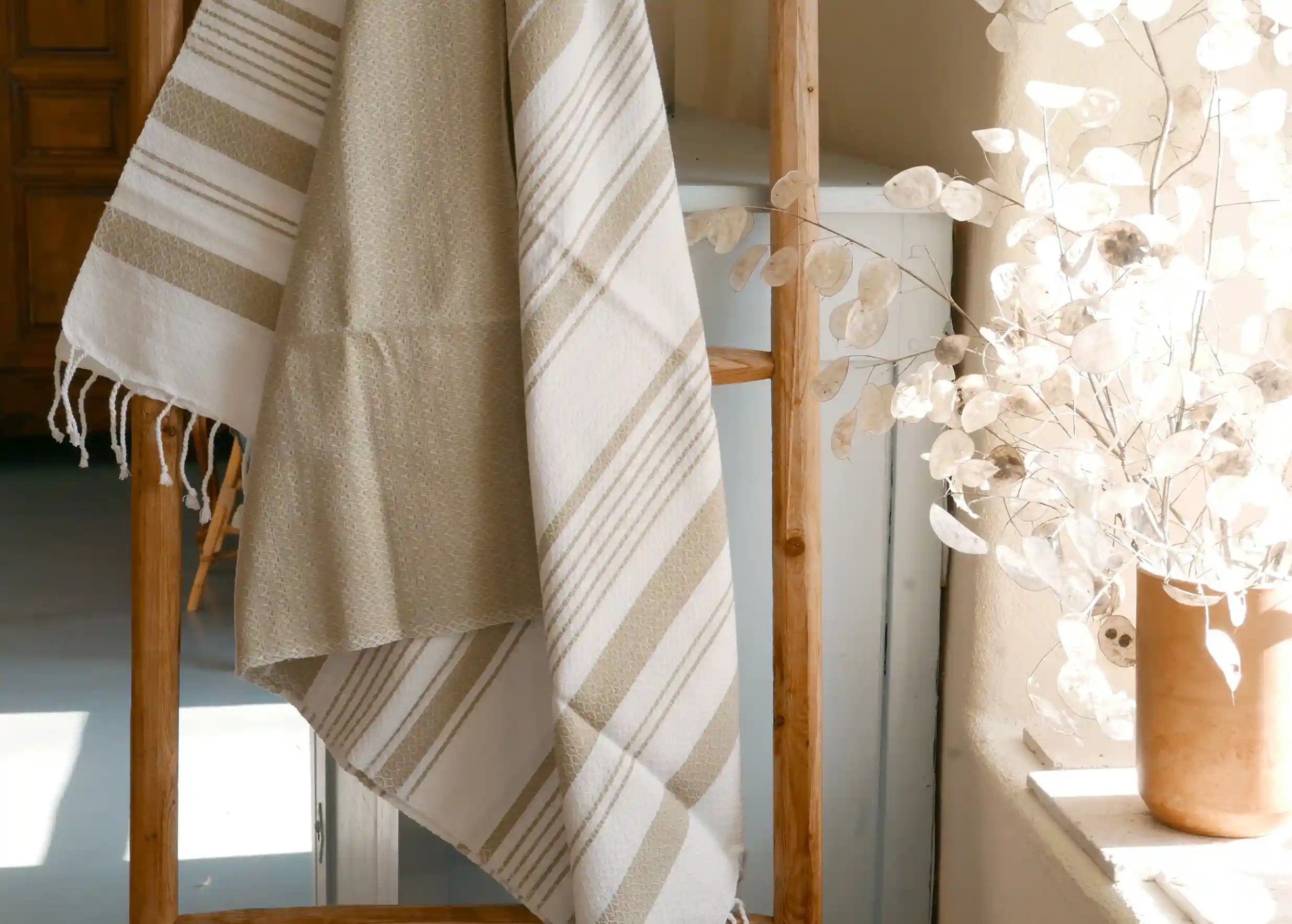 Ginger-coloured fouta hanging near a window, letting in golden light, vase and plant with dried leaves