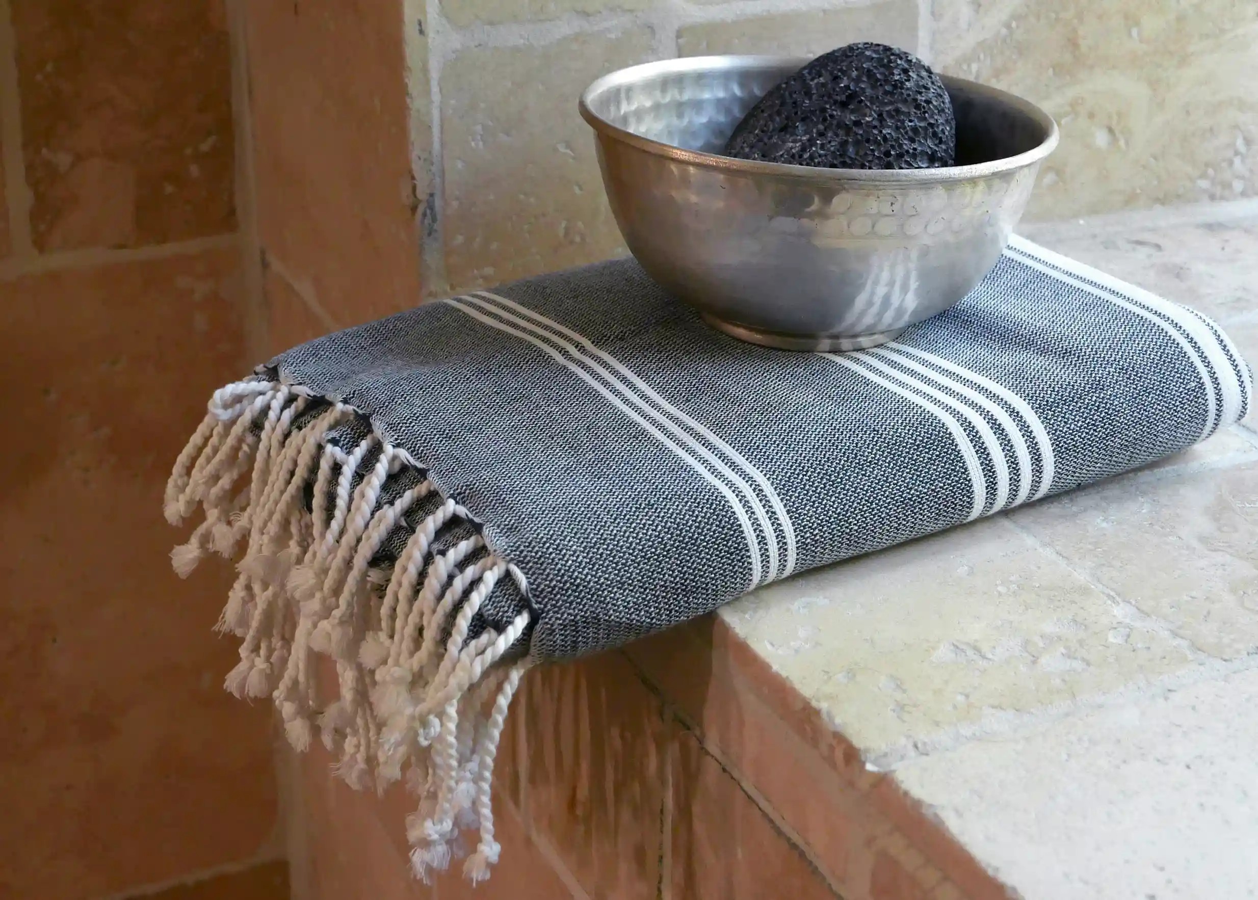 Fouta en coton bio anthracite aux rayures blanches, pliée et posée sur un rebord en pierre, avec un bol en laiton Fouta en coton bio anthracite aux rayures blanches, pliée et posée sur un rebord en pierre, avec un bol en laiton