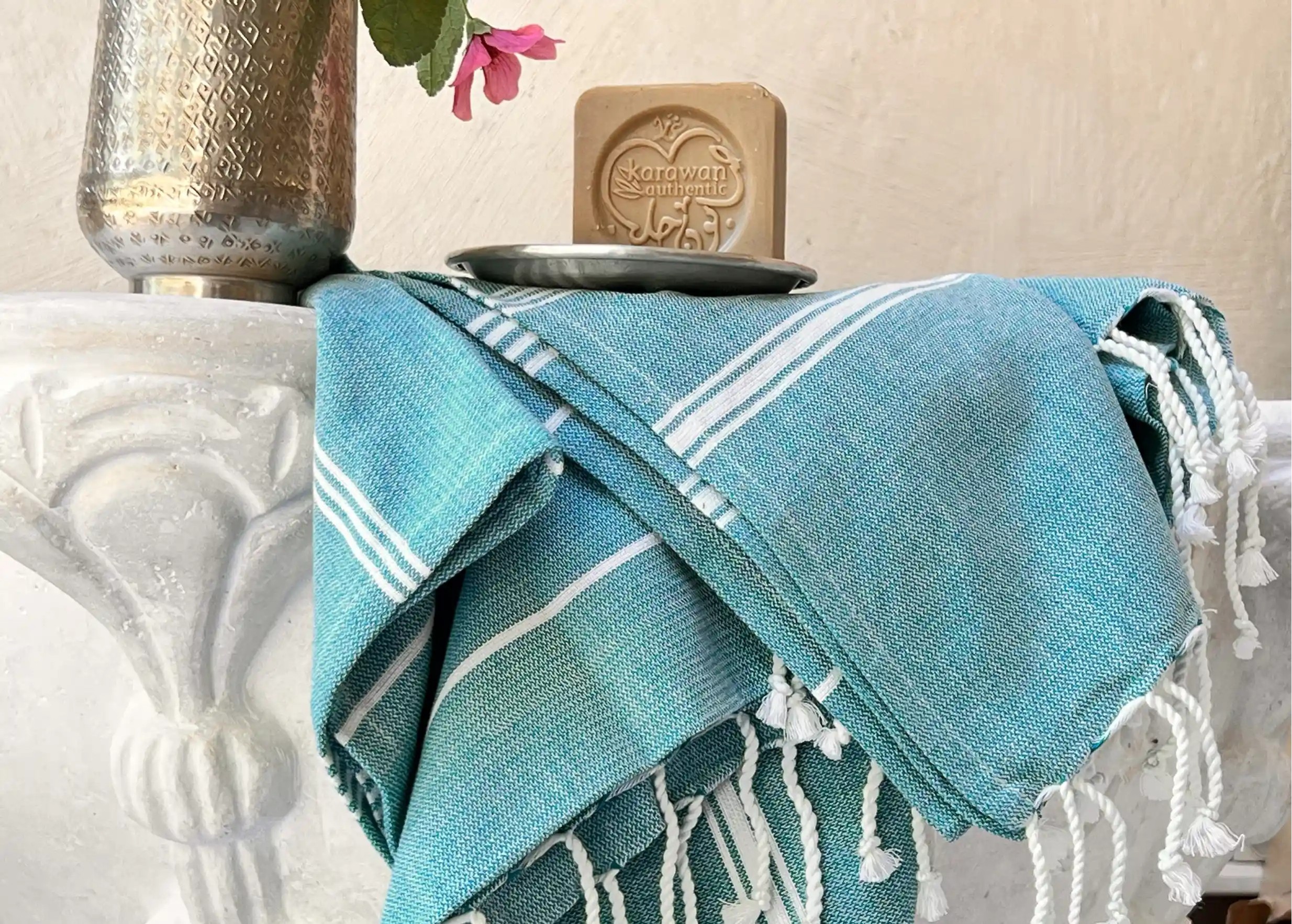 Fouta bleue en coton biologique sur un meuble en pierre, savon d'Alep dans une coupelle en laiton et vase en alu recyclé Fouta bleue en coton biologique sur un meuble en pierre, savon d'Alep dans une coupelle en laiton et vase en alu recyclé