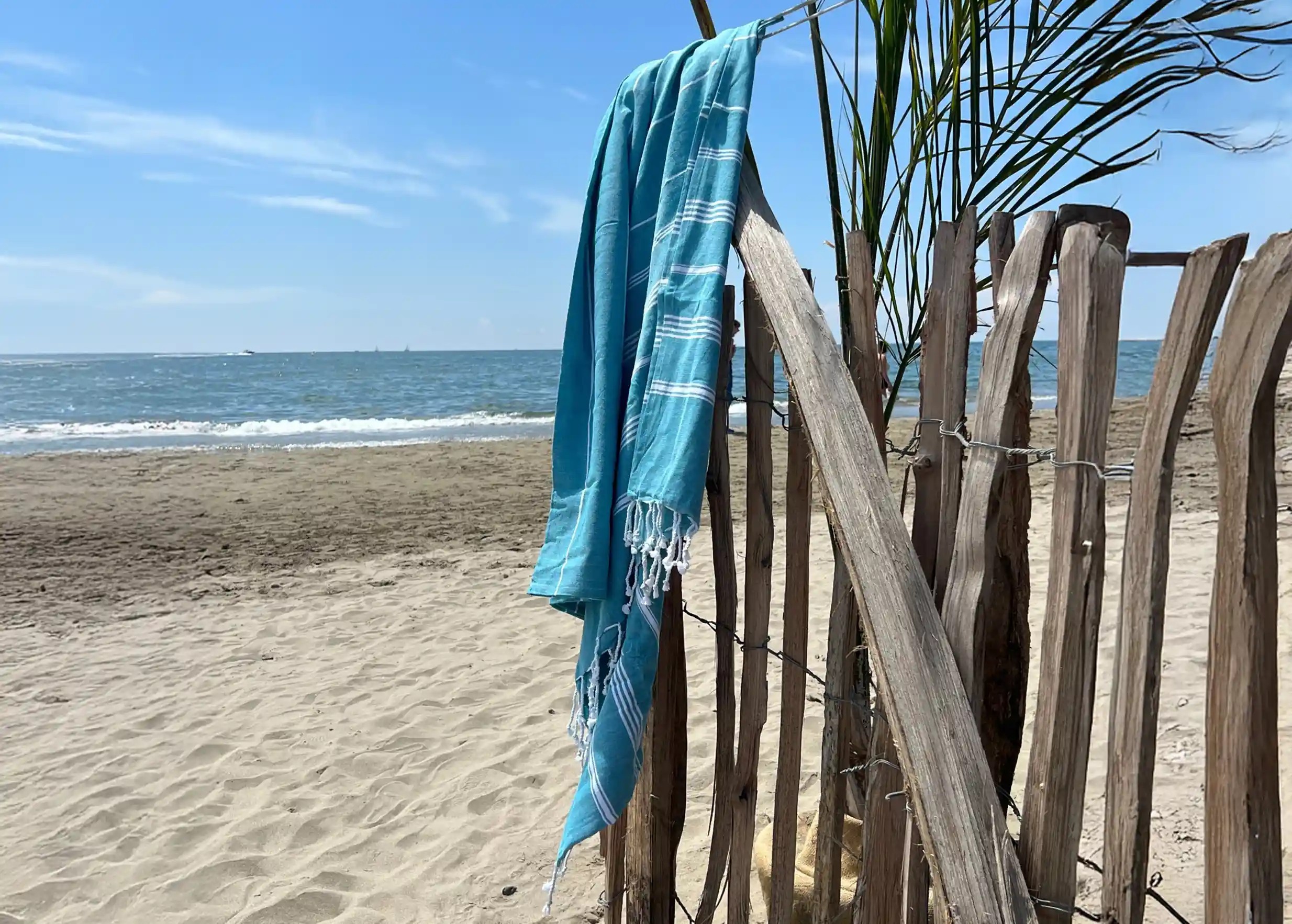 Fouta en coton bio bleu atoll, accrochée à une clôture en bois sur la plage, la mer en arrière-plan Fouta en coton bio bleu atoll, accrochée à une clôture en bois sur la plage, la mer en arrière-plan