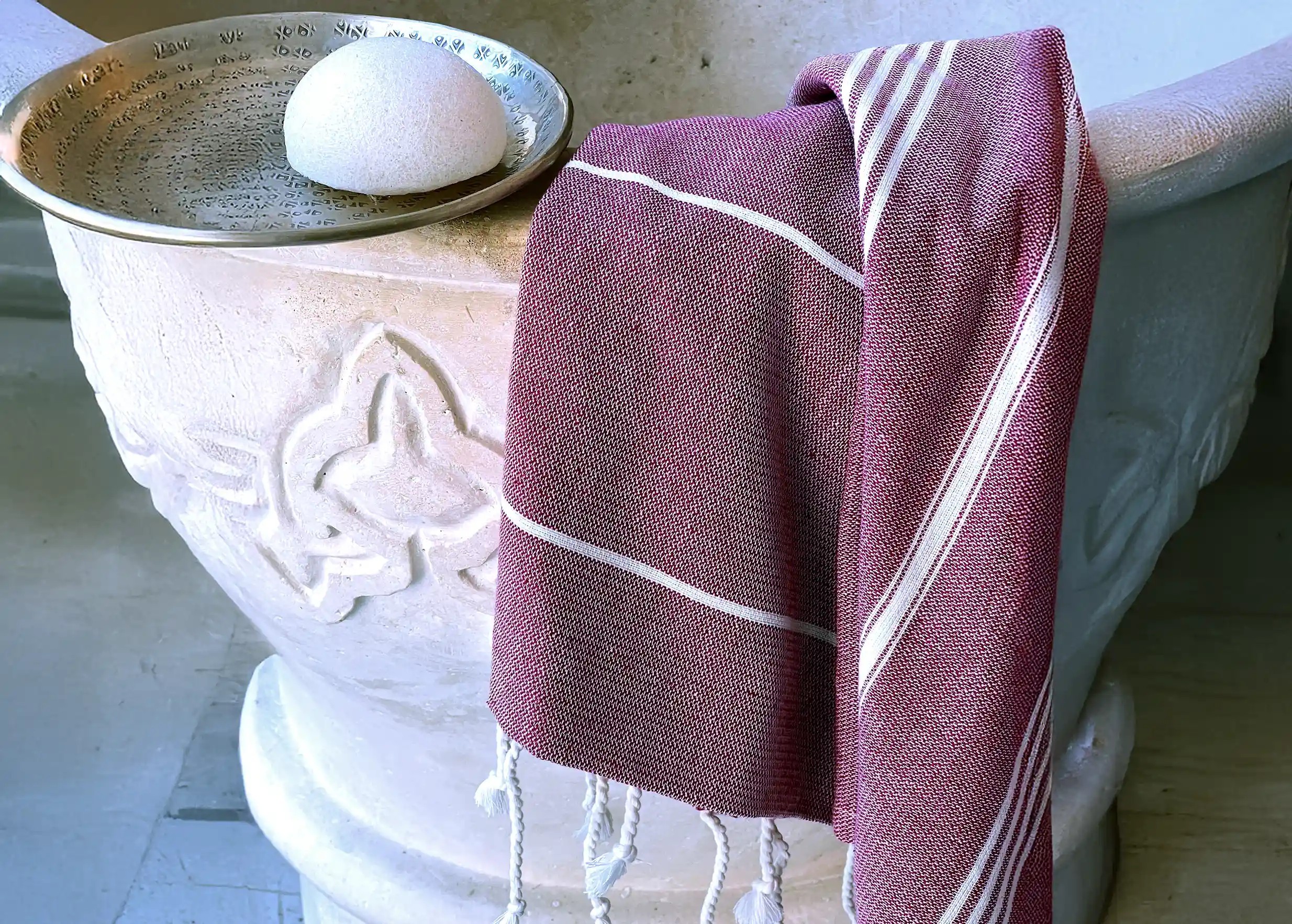 Fouta en coton bio bordeaux, tissée sur chaîne blanche, suspendue à un mur à côté d'une coupelle en laiton Fouta en coton bio bordeaux, tissée sur chaîne blanche, suspendue à un mur à côté d'une coupelle en laiton