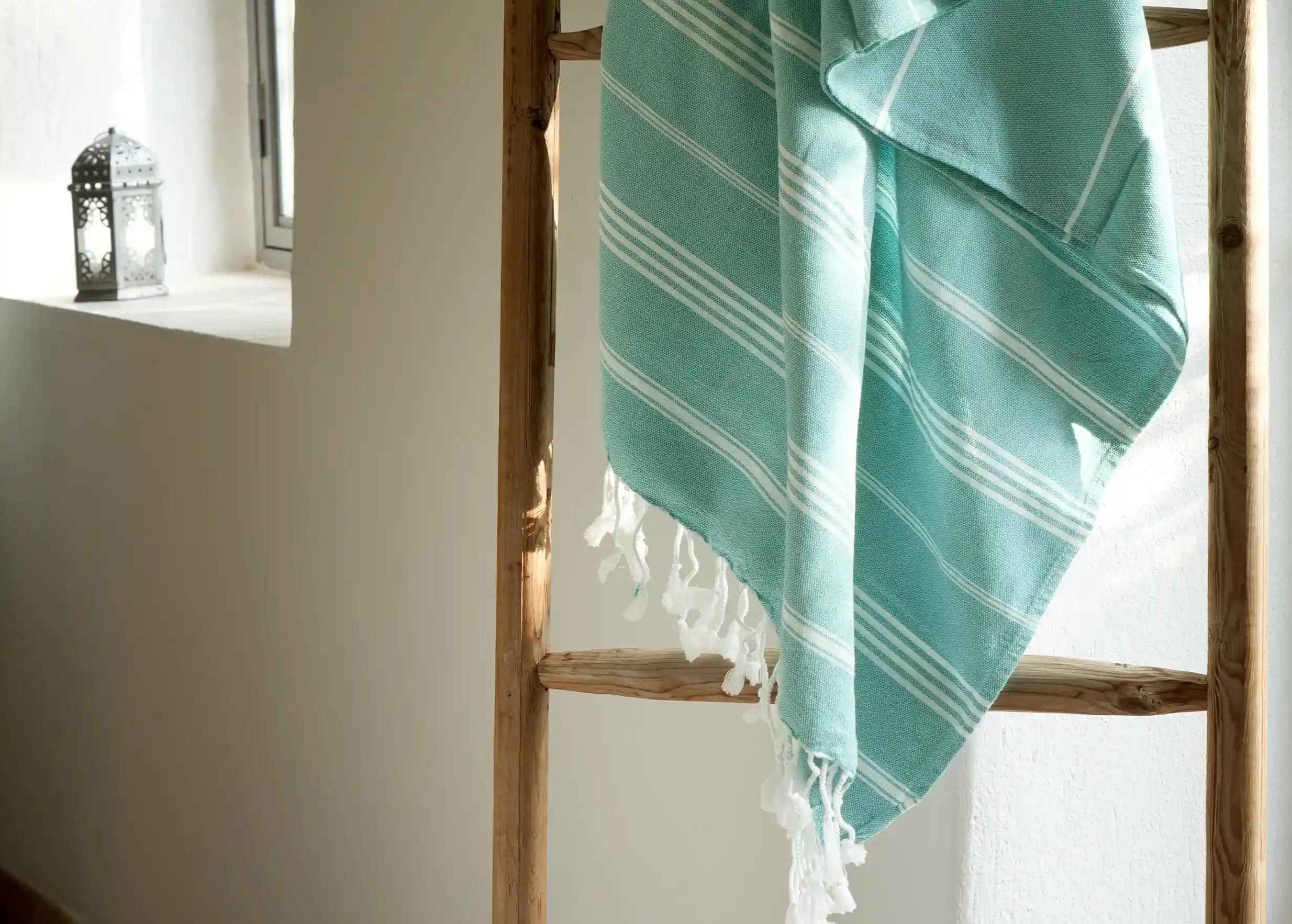 Fouta menthe avec rayures blanches et franges, accrochée à une échelle en bois, dans un intérieur lumineux et minimaliste Fouta menthe avec rayures blanches et franges, accrochée à une échelle en bois, dans un intérieur lumineux et minimaliste