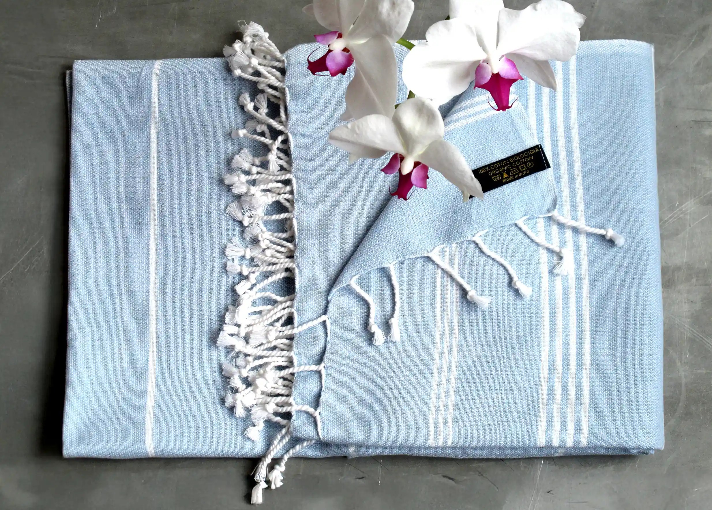Fouta en coton bio au ton bleu clair, des rayures blanches et des franges nouées, pliée avec une fleur d'orchidée posée dessus Fouta en coton bio au ton bleu clair, des rayures blanches et des franges nouées, pliée avec une fleur d'orchidée posée dessus