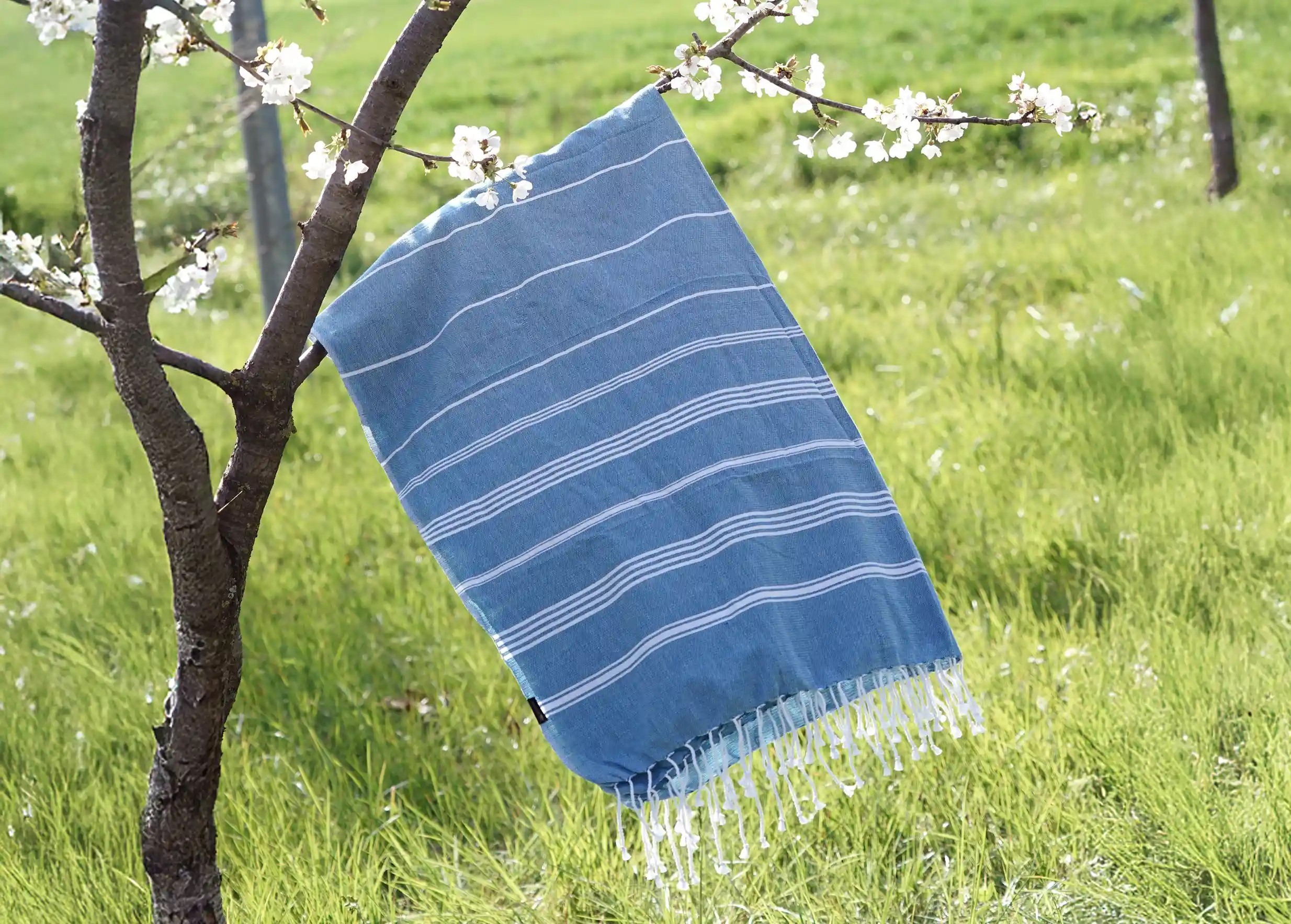 Fouta en coton biologique GOTS, coloris océan, flottant au vent près d'une branche d'arbre en fleurs sous un ciel bleu Fouta en coton biologique GOTS, coloris océan, flottant au vent près d'une branche d'arbre en fleurs sous un ciel bleu