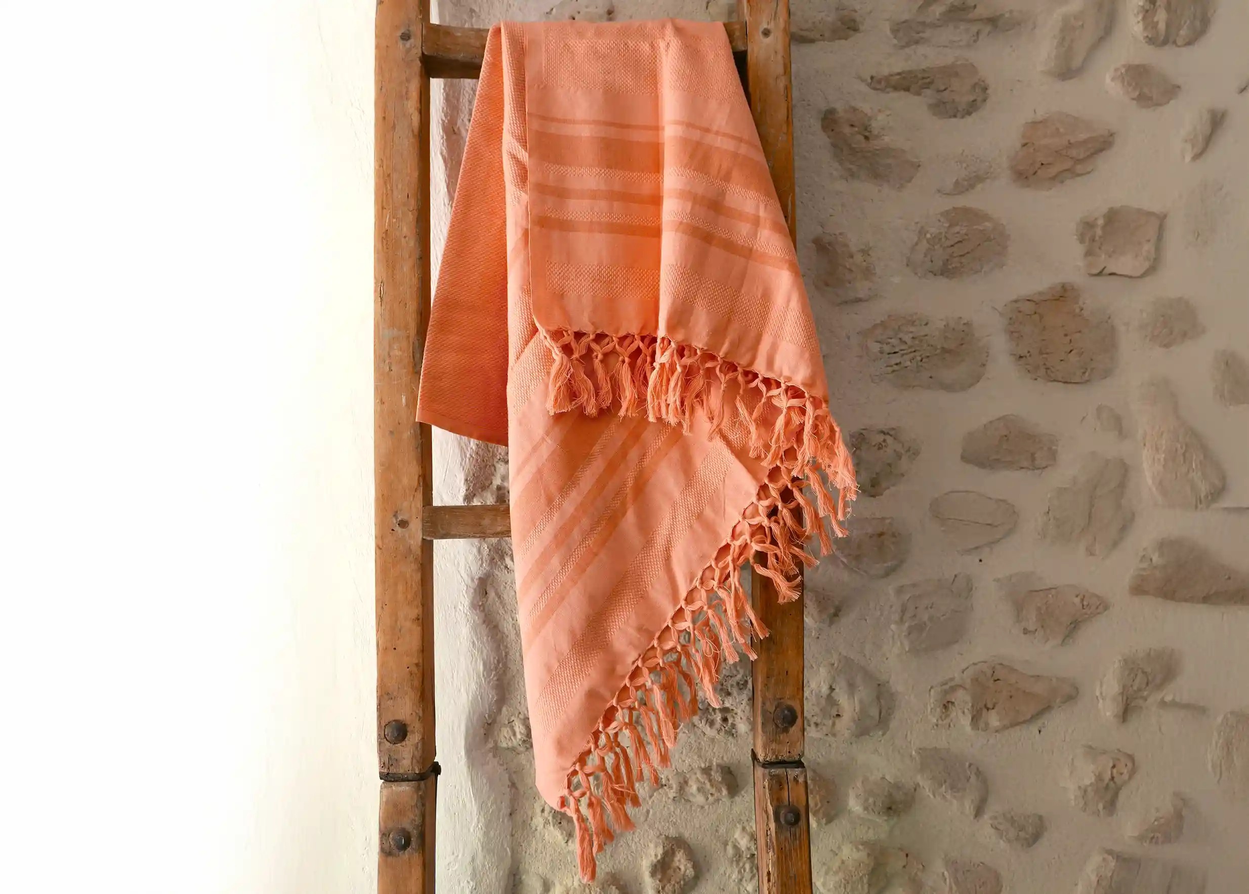 Coral-coloured fouta in organic cotton with honeycomb weave against wooden ladder, stone wall