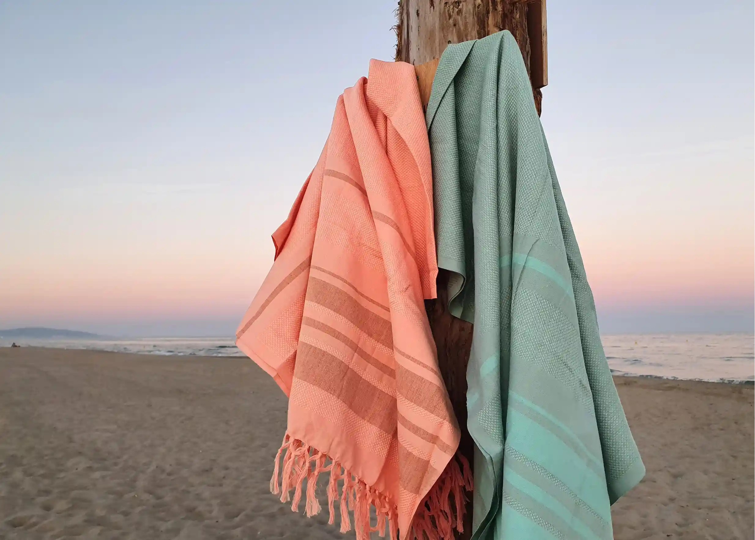 Coral and blue coloured foutas hanging from a post on the beach, alternating two-tone stripes
