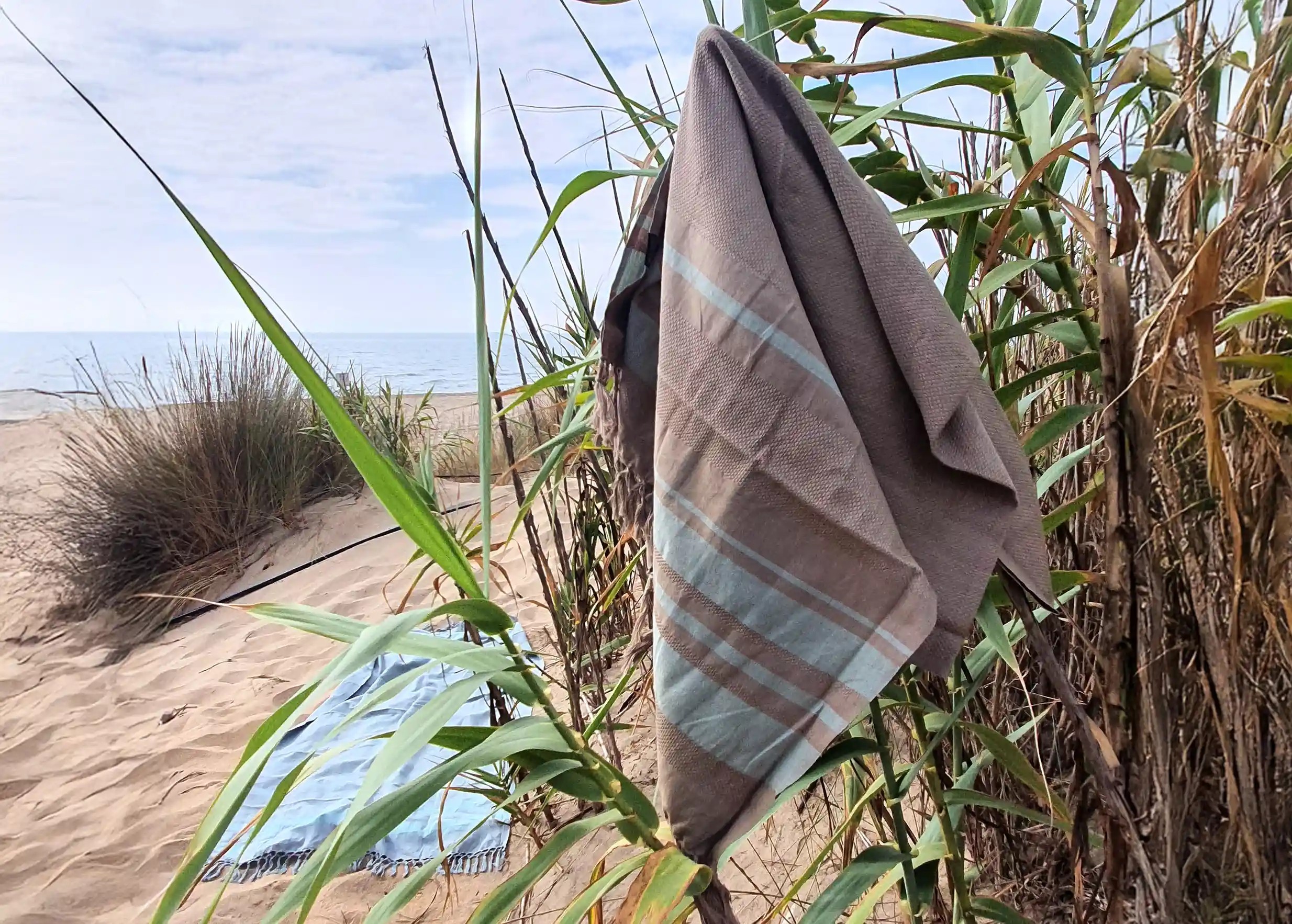 Earth beige organic cotton fouta suspended and floating lightly in the wind with a sandy beach and the sea in the background