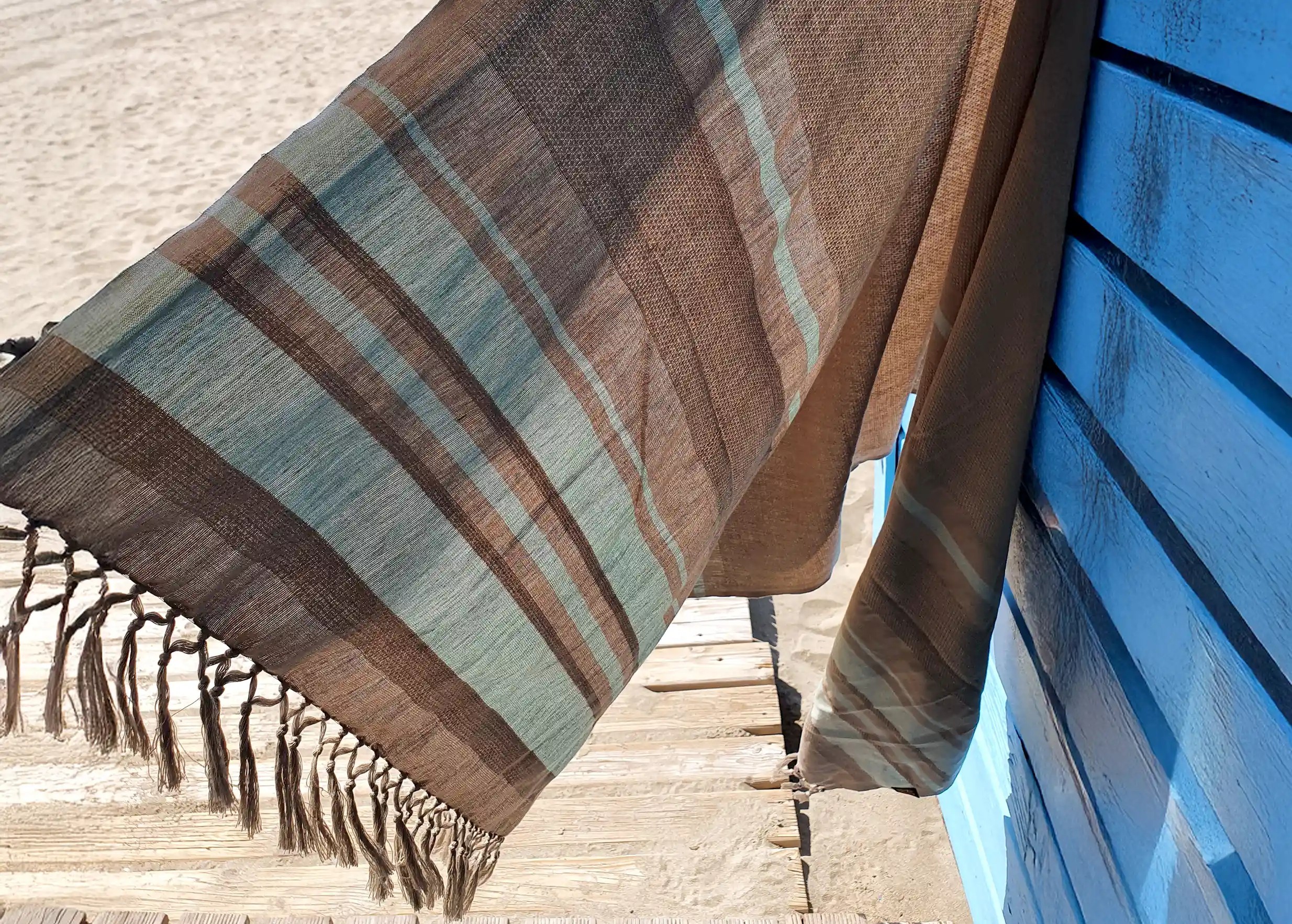 Close-up of an earth beige fouta in organic cotton with brown and blue stripes, beach and light blue sky in the background