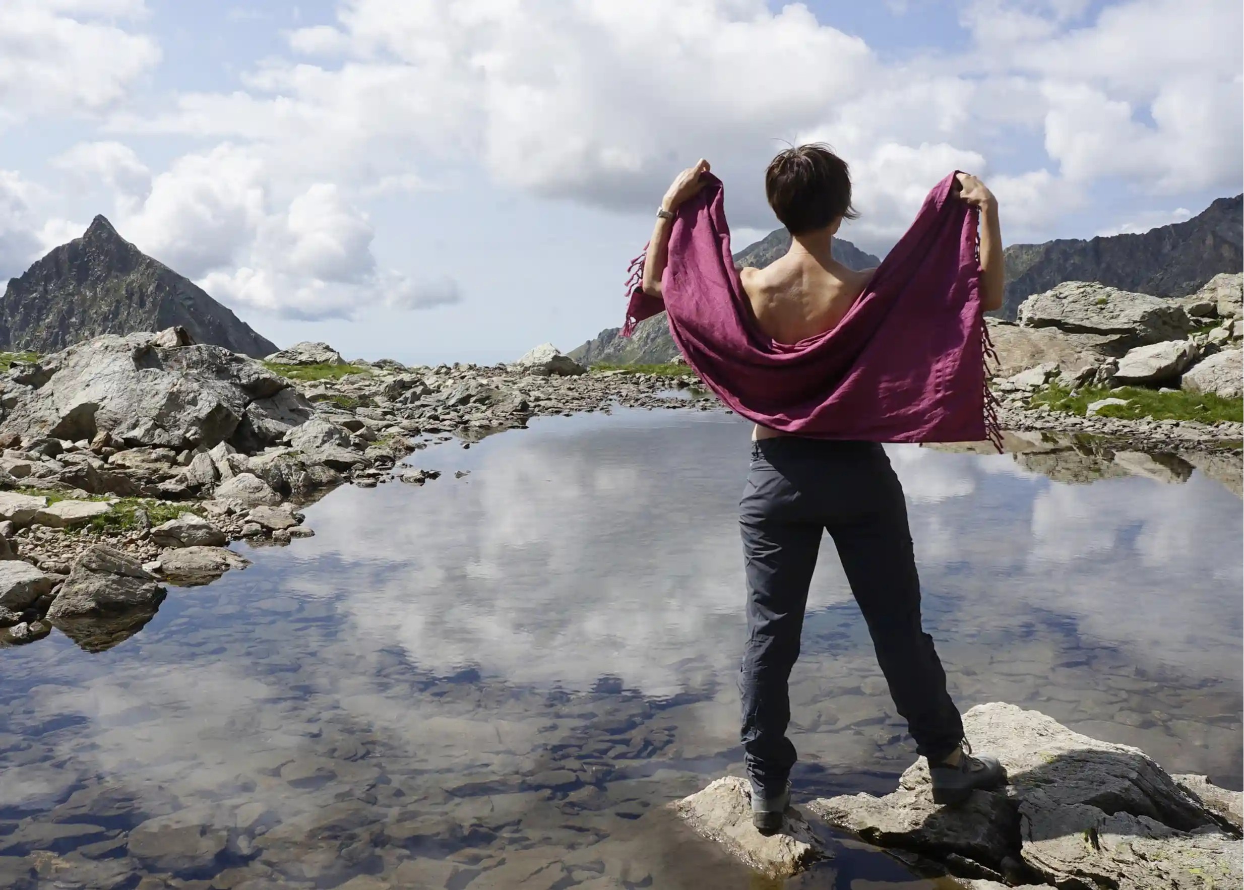 Personne debout de dos, tenant une fouta en coton bio fushia devant un paysage de montagne rocheux avec un lac