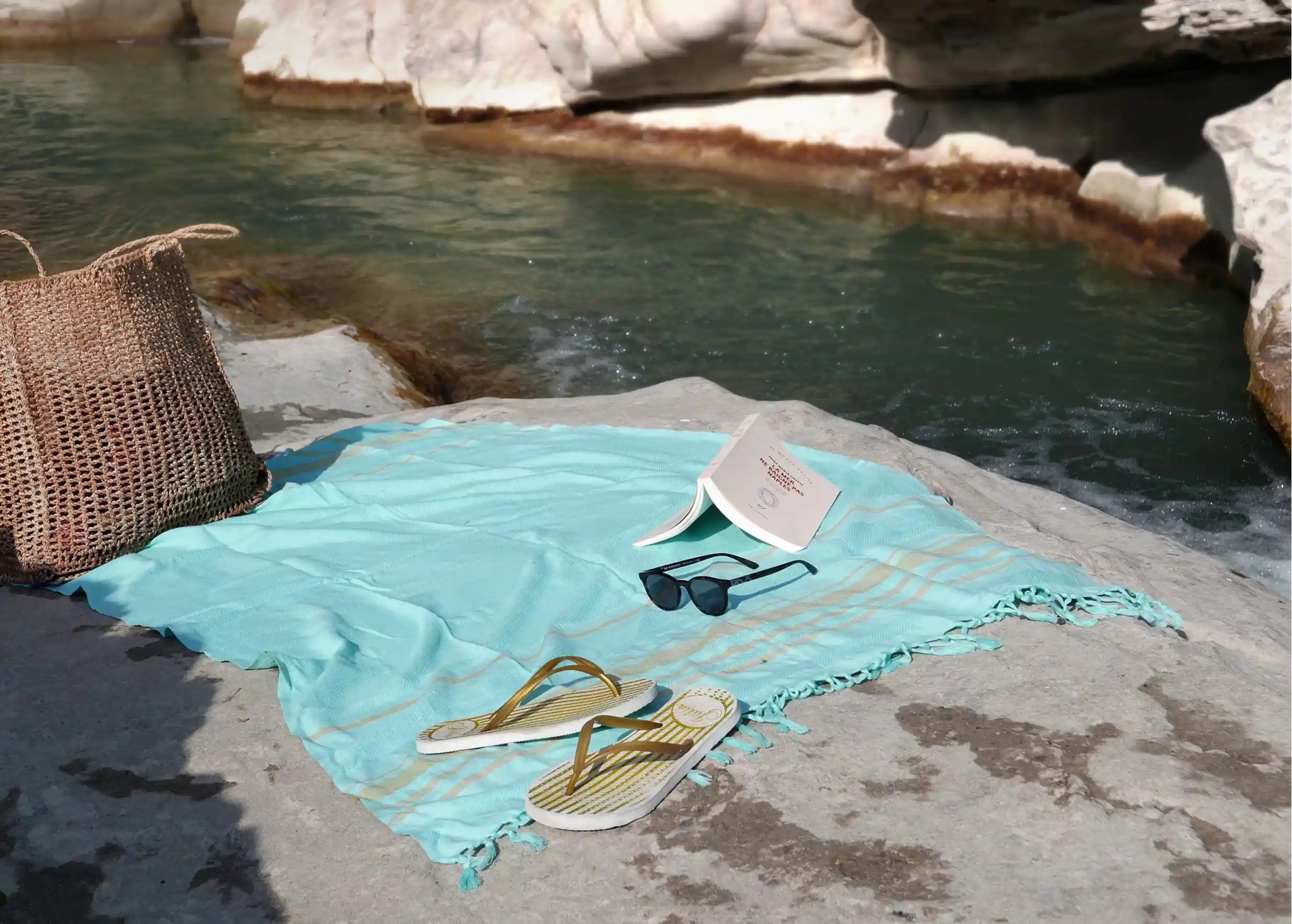 Fouta en coton biologique turquoise étalée sur un rocher près de l'eau, avec un livre posé dessus et un panier en osier
