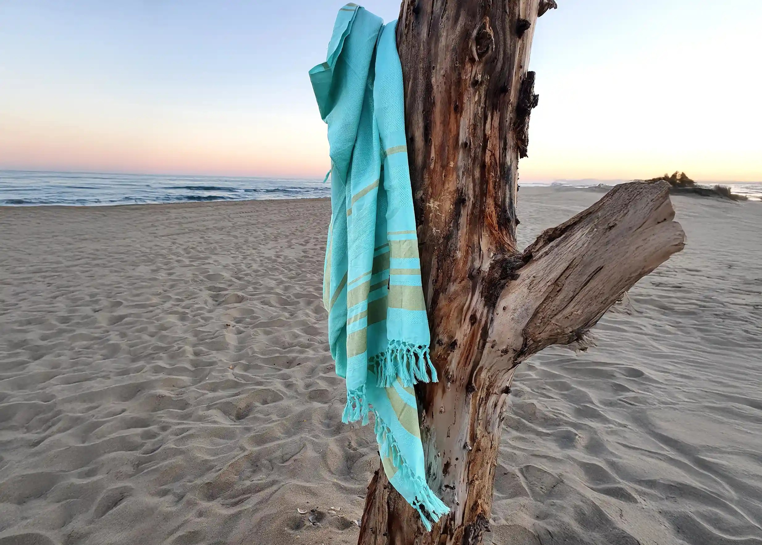 Turquoise-coloured fouta in striped organic cotton, hanging from a branch outdoors, green vegetation