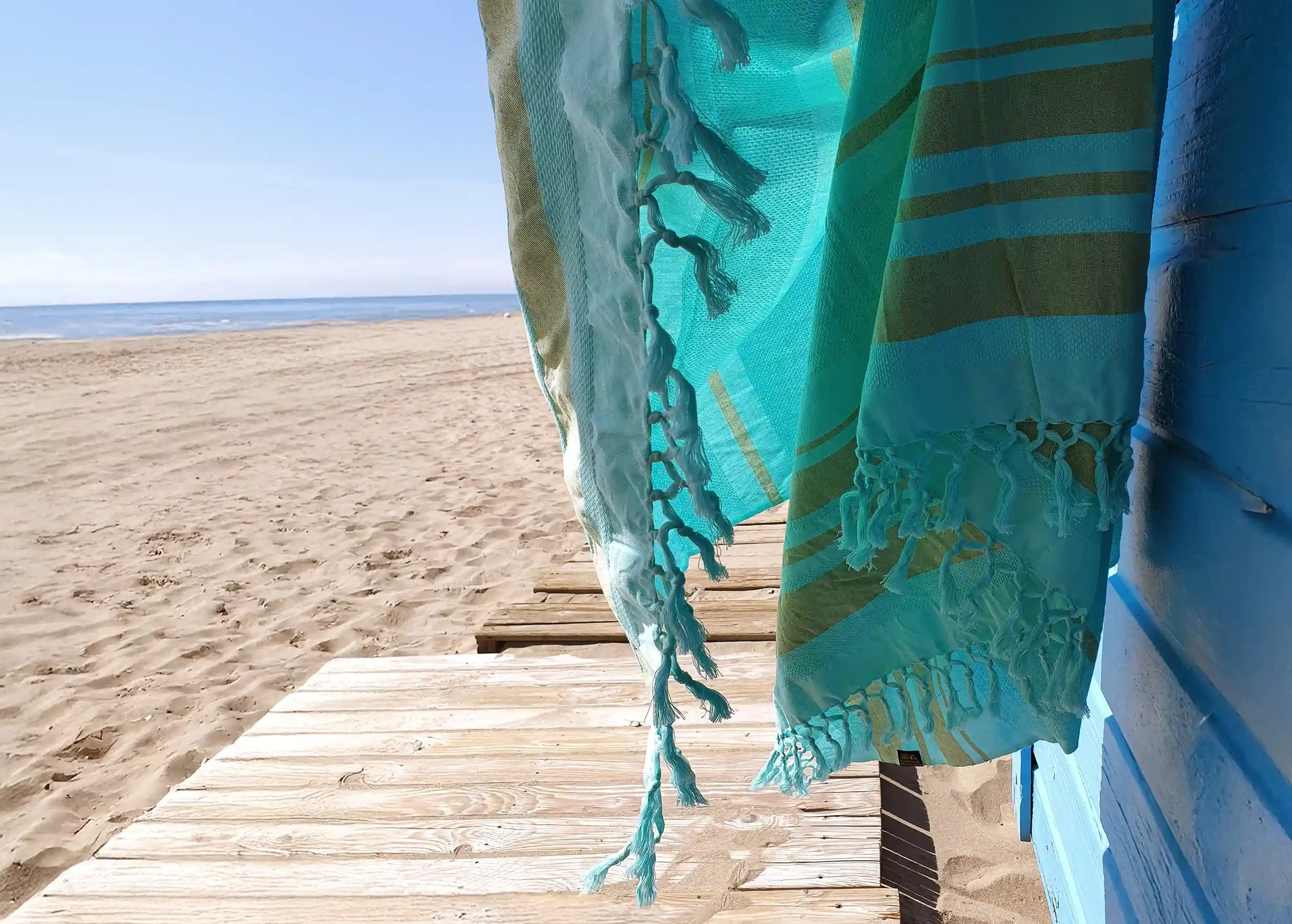 Close-up of turquoise-coloured organic cotton fouta with turquoise fringes, beach and sea in the distance