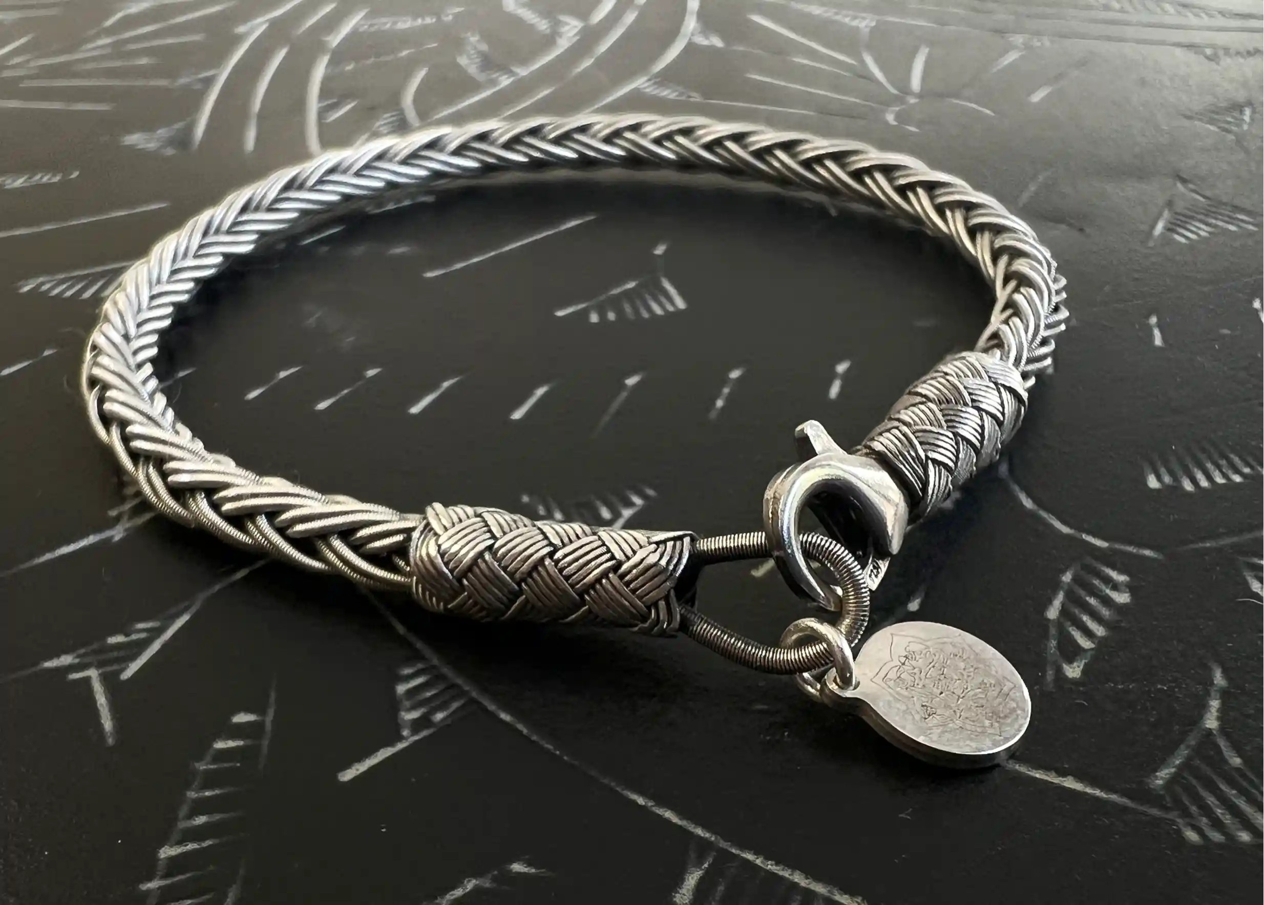 Kokaz braided silver bracelet in round form with metal clasp and small engraved medal, set on a black surface
