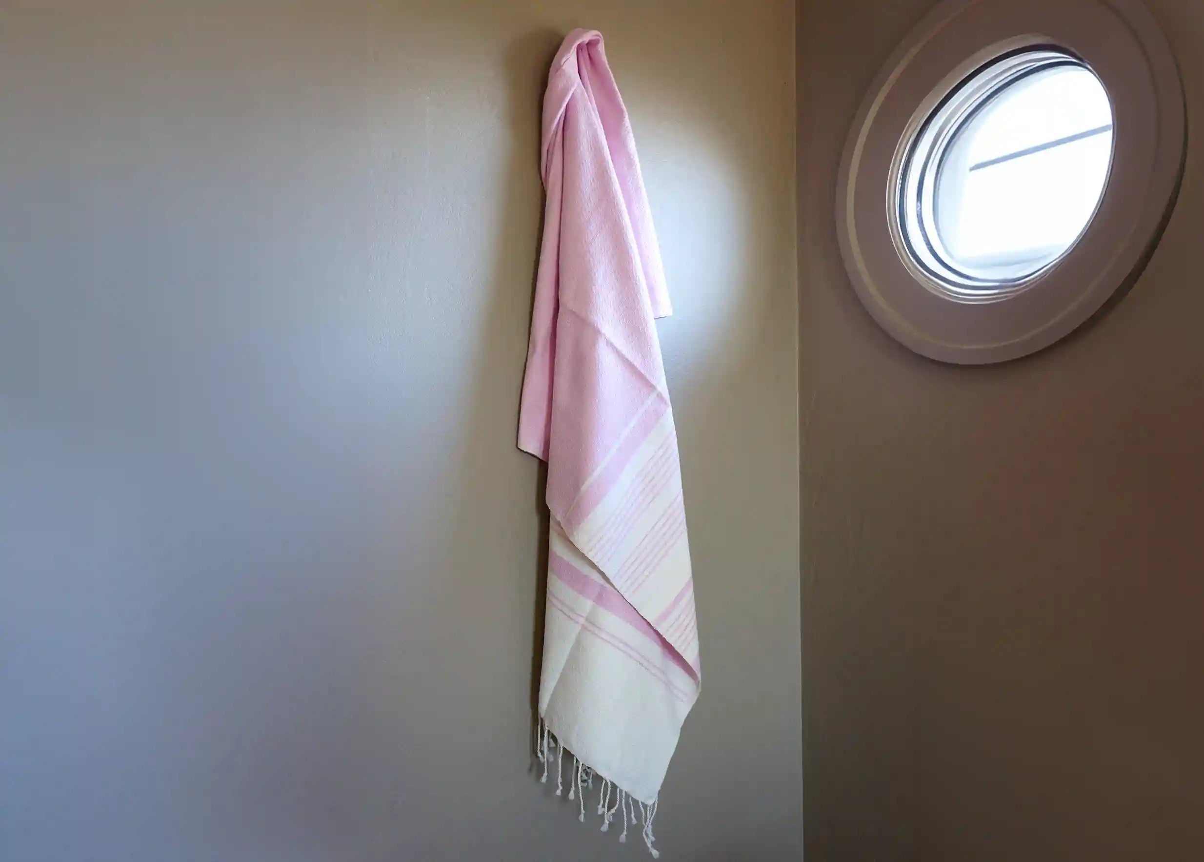 Une fouta en coton bio de couleur parme par Karawan, suspendue contre un mur, à côté d'une petite fenêtre ronde de style hublot