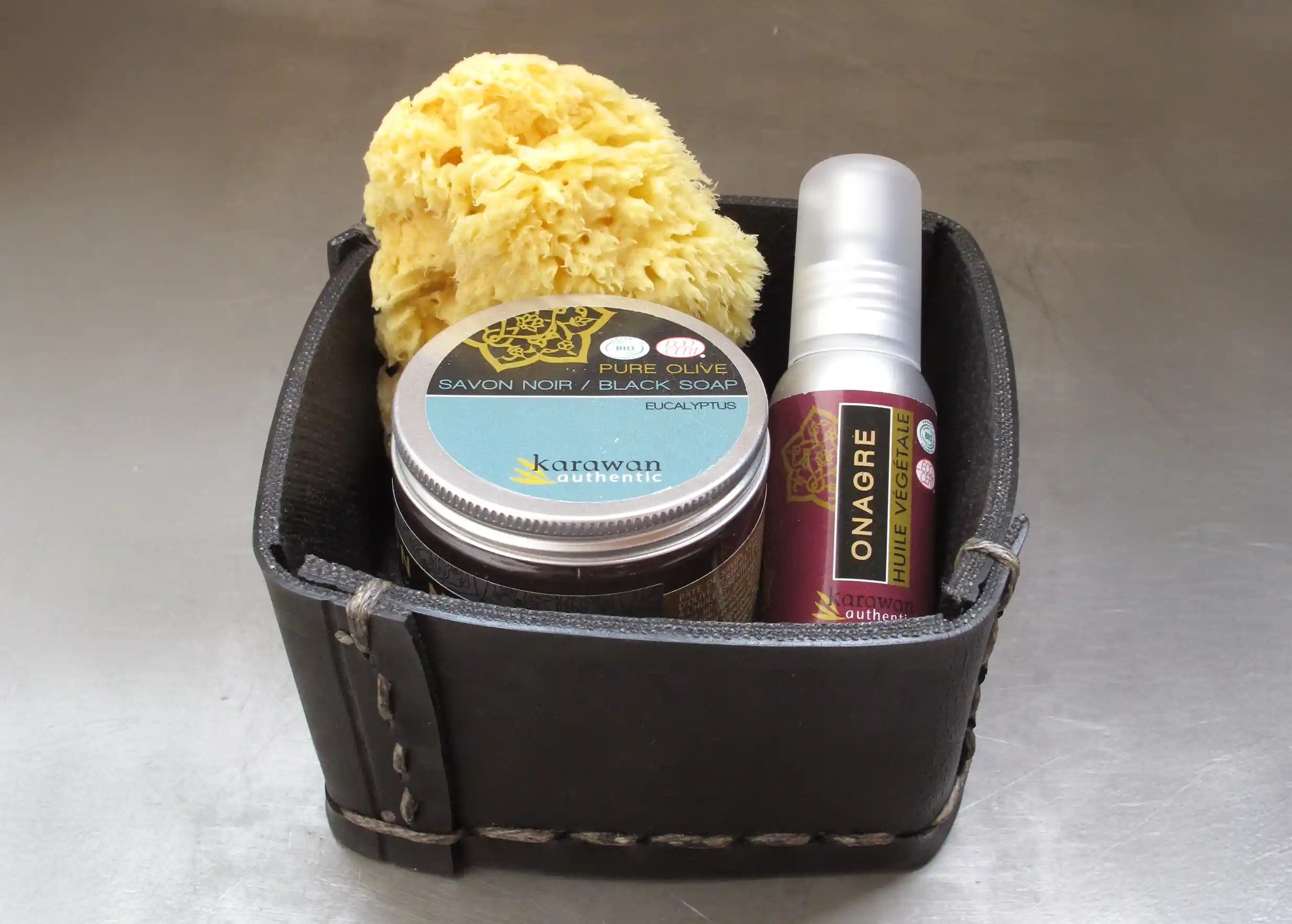 Small square box made from recycled tyres, containing a jar of black soap, a natural sponge and a bottle of primrose oil.