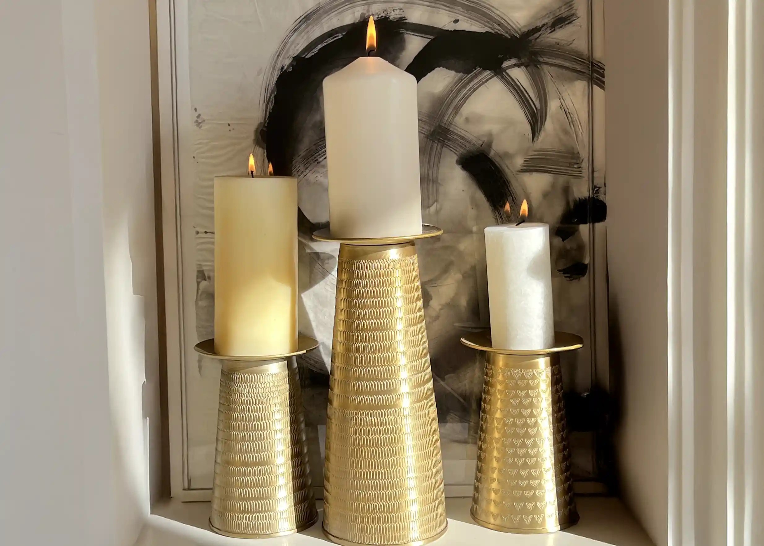 Un bougeoir doré en alu et texturé, portant une bougie allumée, une œuvre d'art colorée en arrière-plan