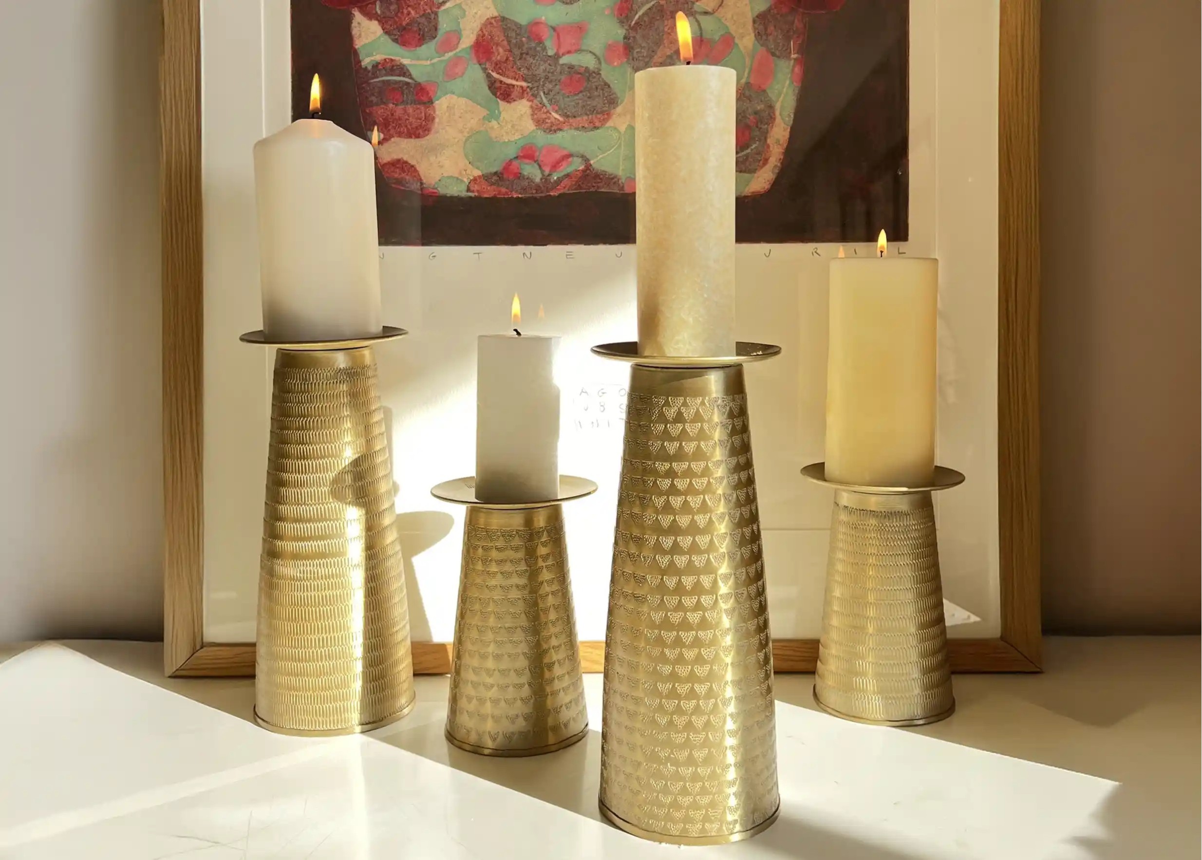 Quatre bougeoirs doré en alu et texturé, portant des bougies allumées, une œuvre d'art colorée en arrière-plan