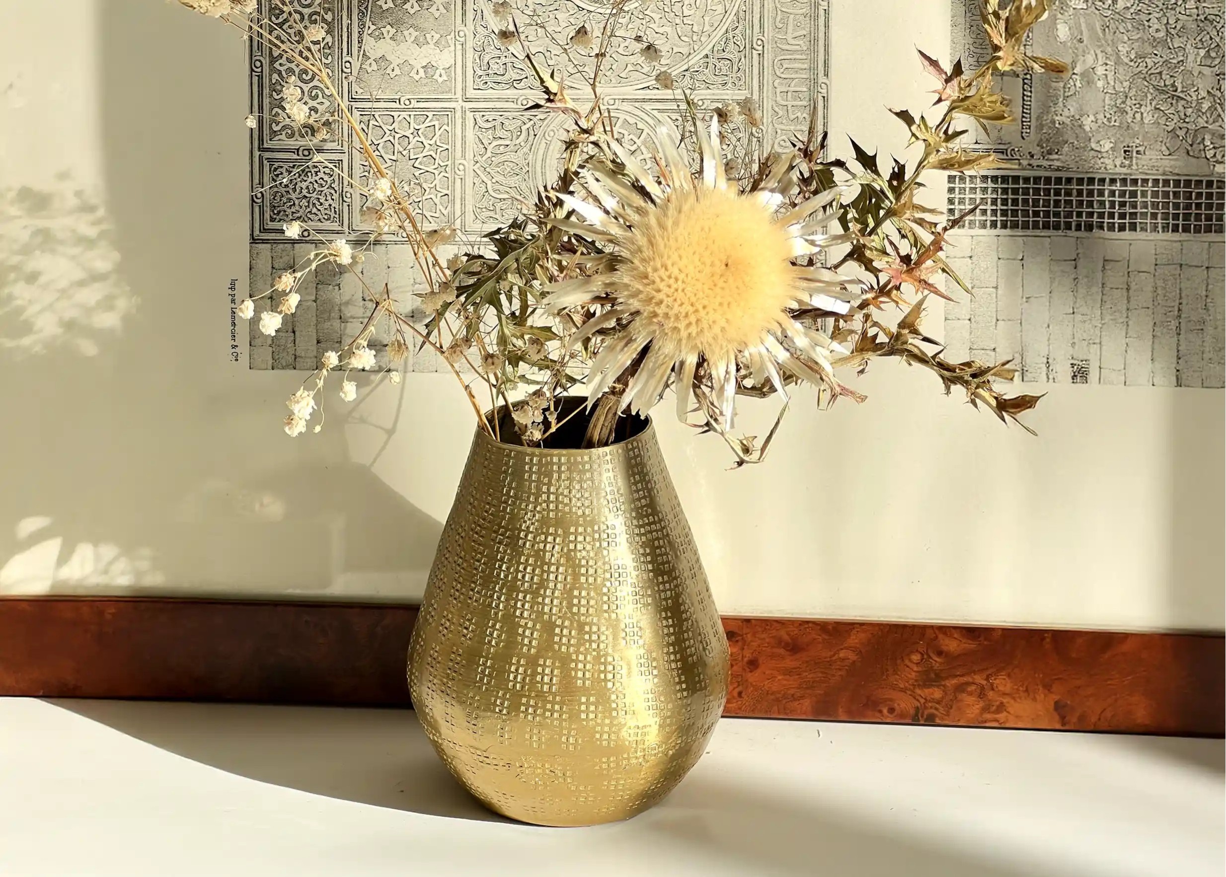 Petit vase en alu avec des fleurs séchées posé devant un cadre décoratif