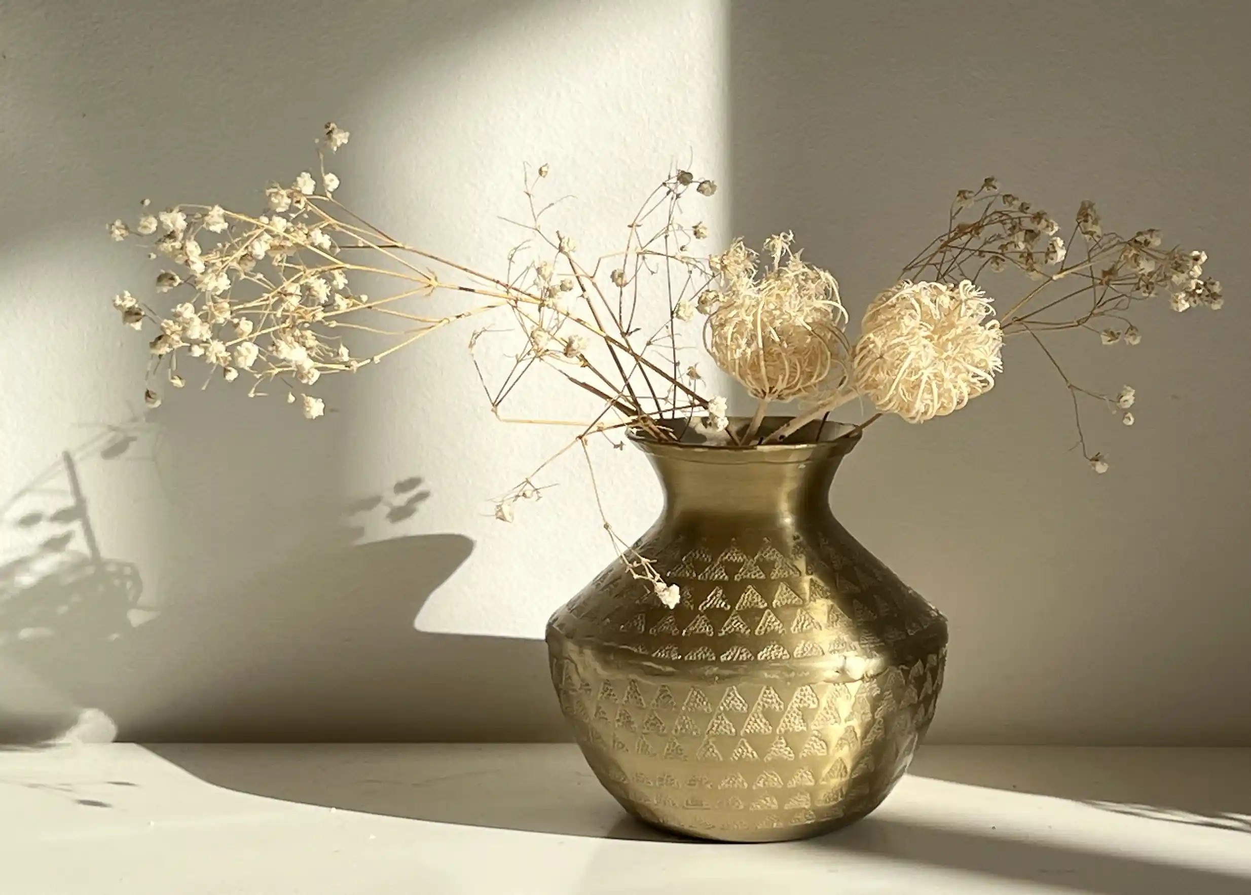 Vase en aluminium recyclé avec une finition dorée, contient des fleurs séchées, ombre projetée sur le mur