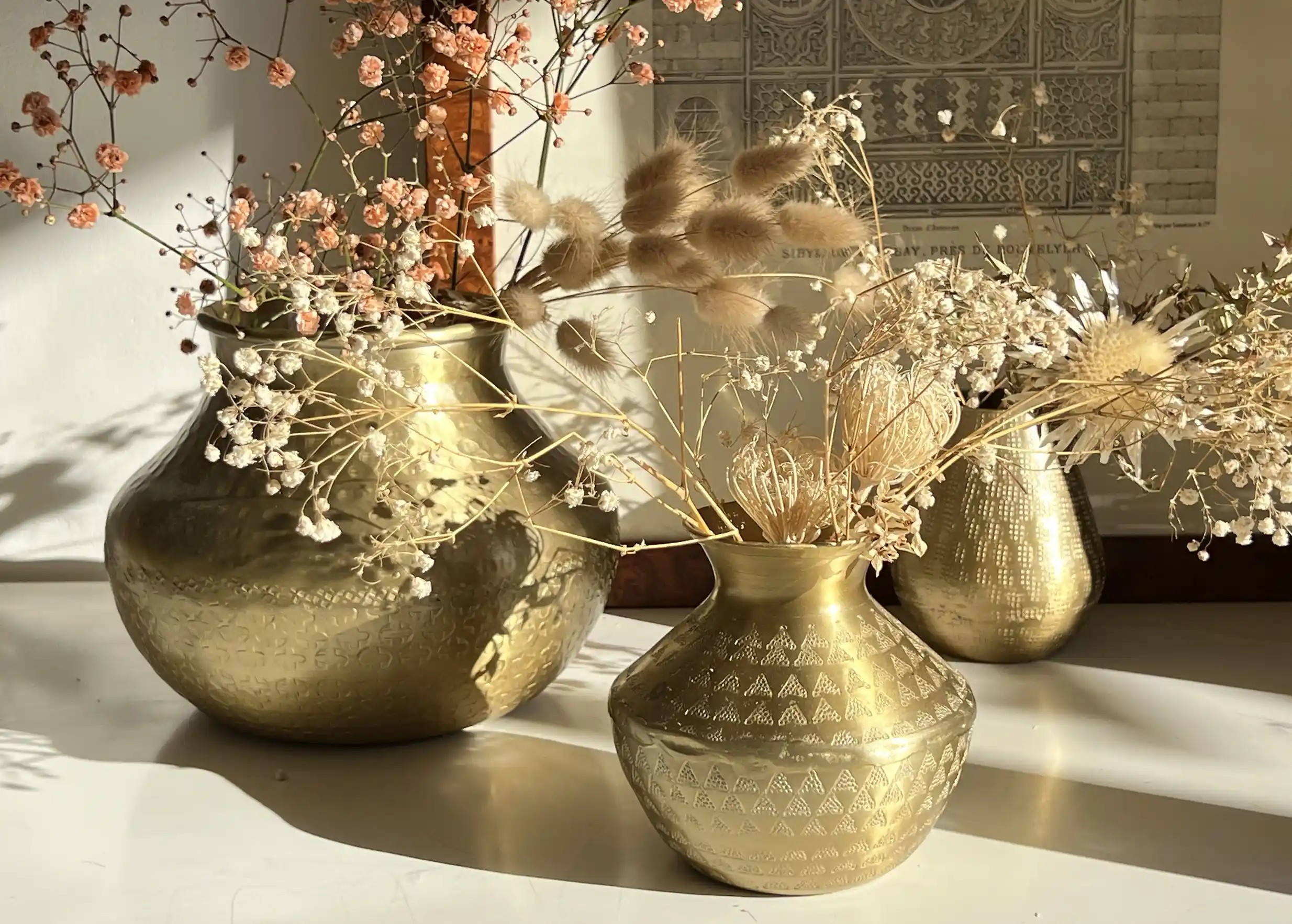 Ensemble de vases, petit vase en alu aux motifs gravés, et grand vase avec fleurs séchées, devant un cadre décoratif