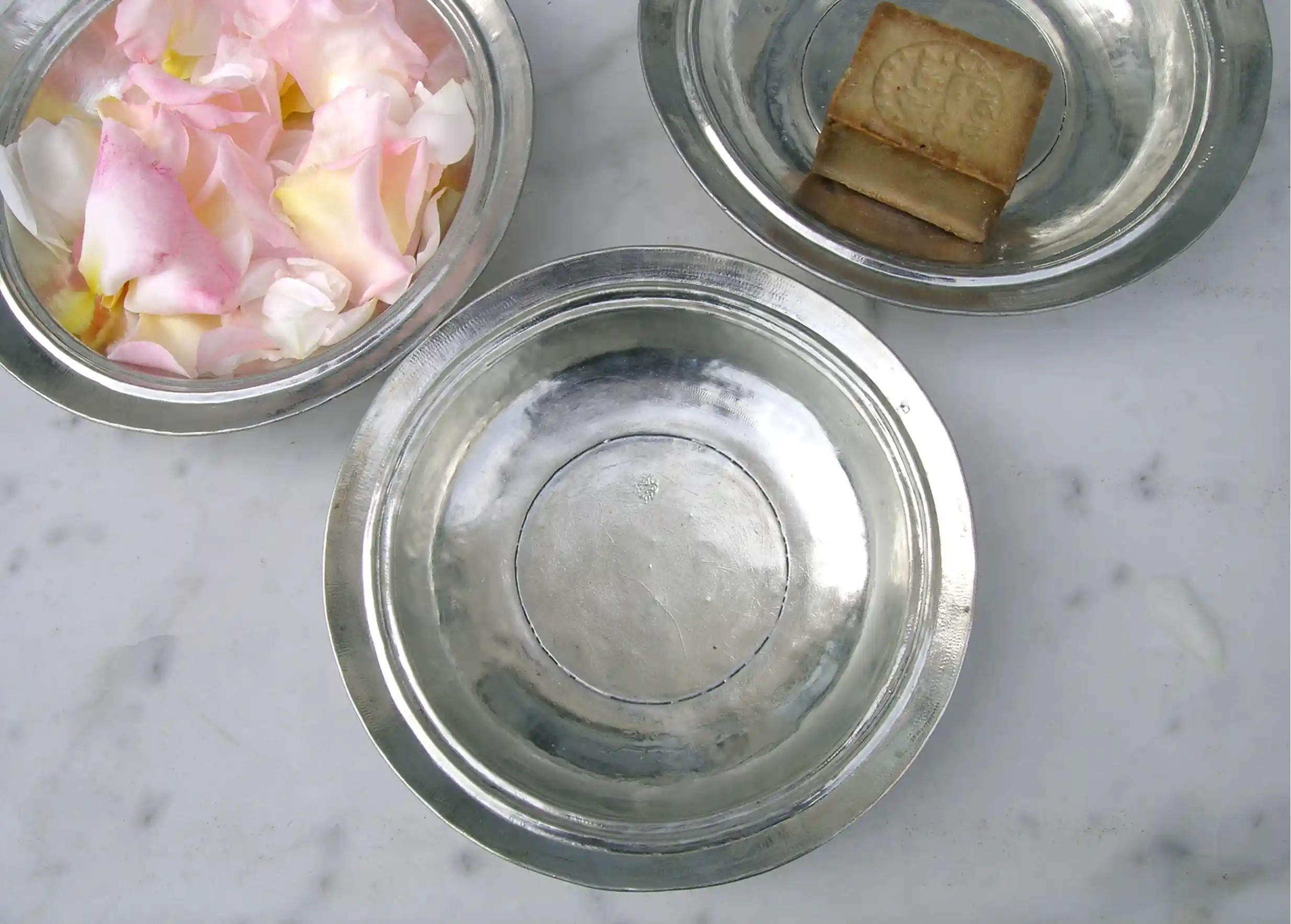 Trois assiettes creuses en cuivre étamé, contenant respectivement des pétales de rose colorés et un savon d'Alep