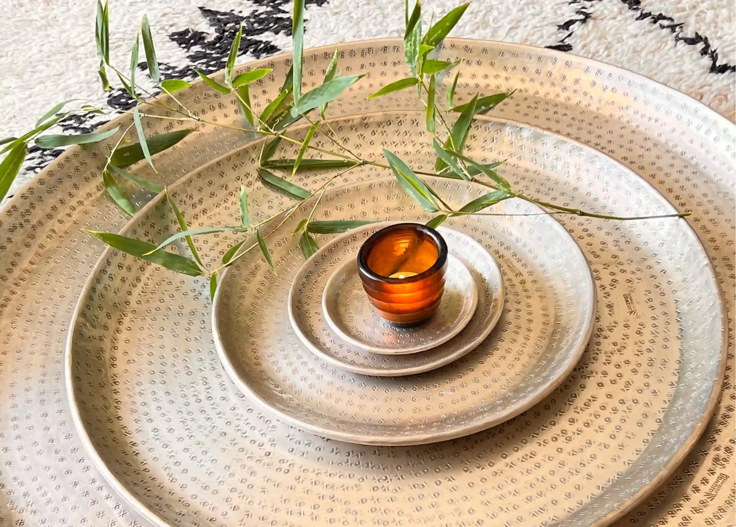 Plateaux en laiton argenté martelé et gravé, posés sur un tapis à motifs noirs et blancs, un photophore avec une bougie allumée