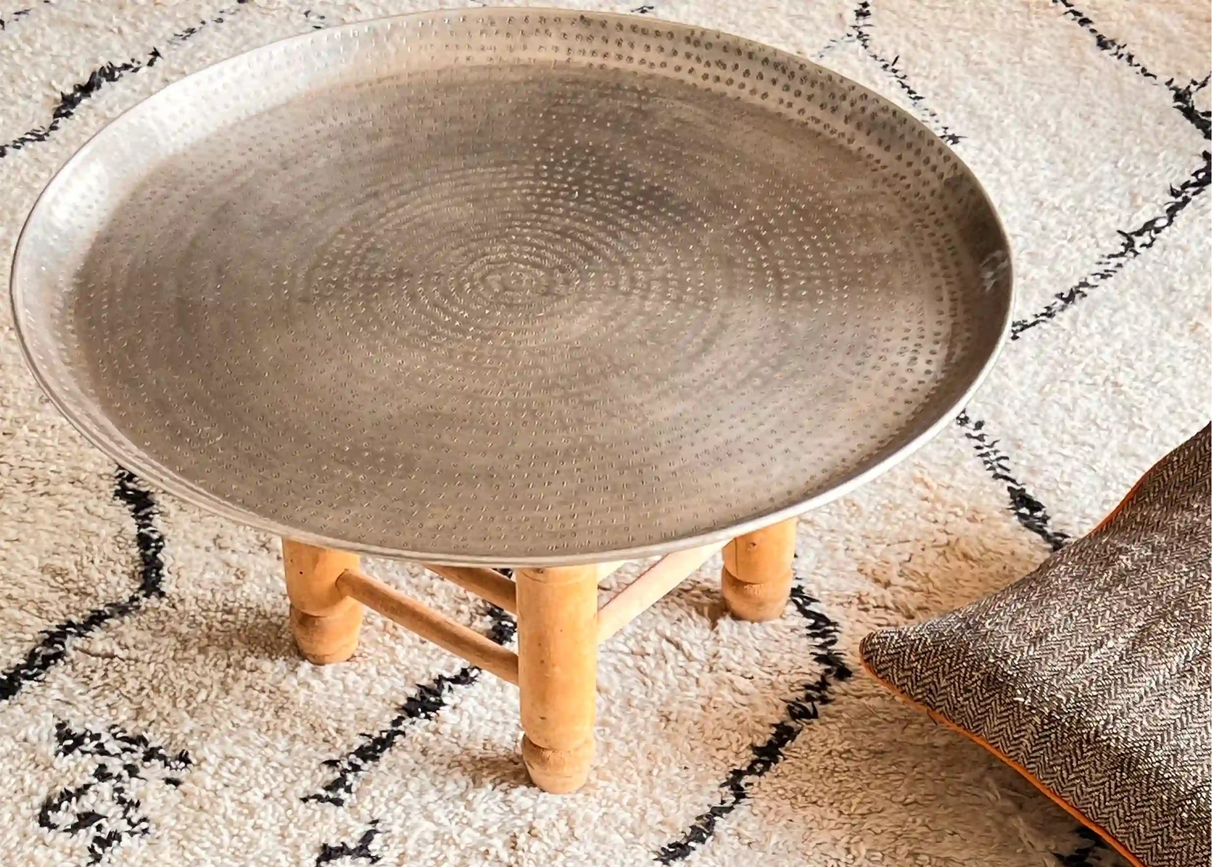 Plateau en laiton argenté martelé et gravé Palmyr posé sur un tabouret en bois, un coussin déco dépassant de la photo