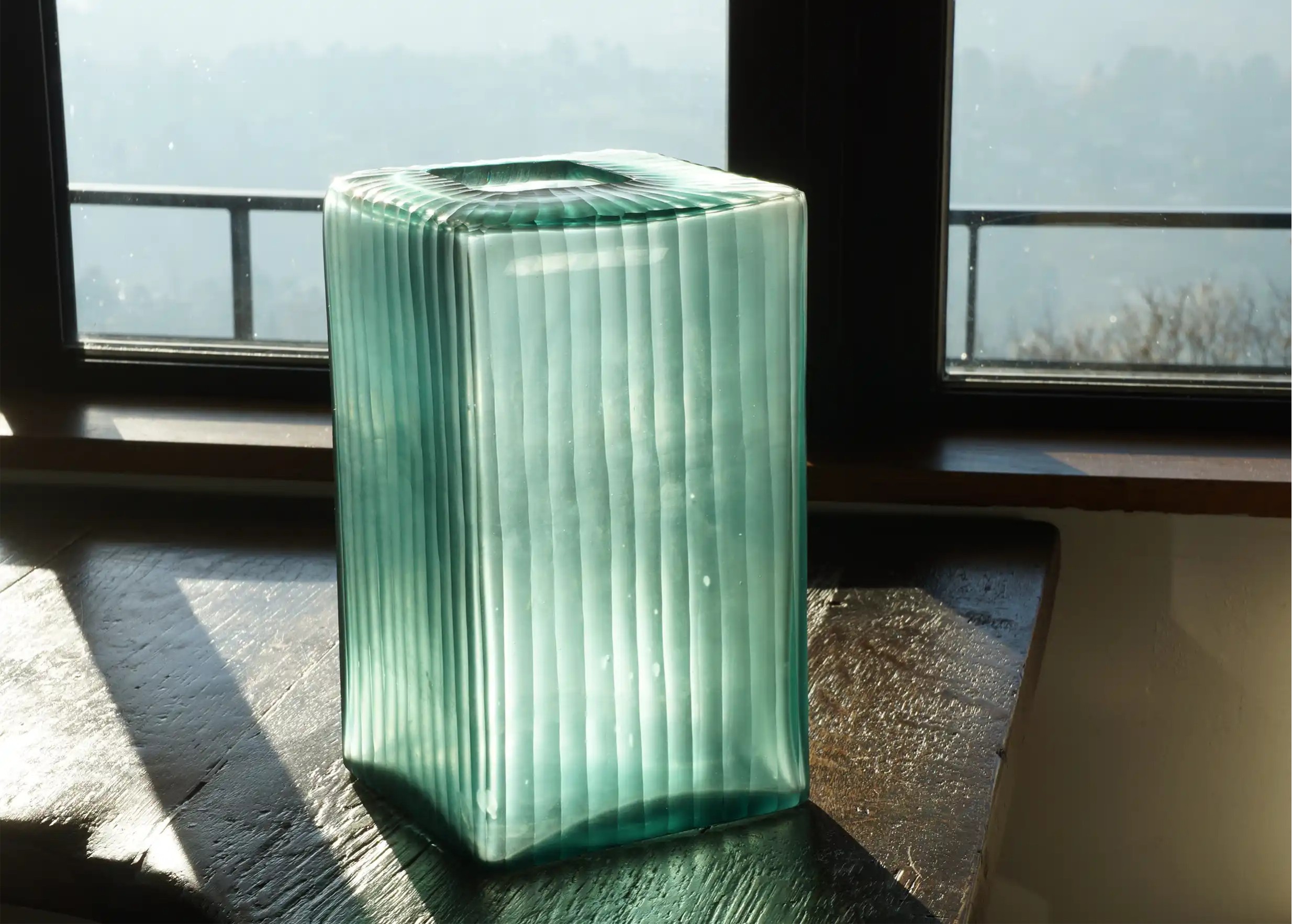 Vase transparent en verre soufflé et recyclé, teinte vert aqua, posé sur une table en bois près d'une fenêtre