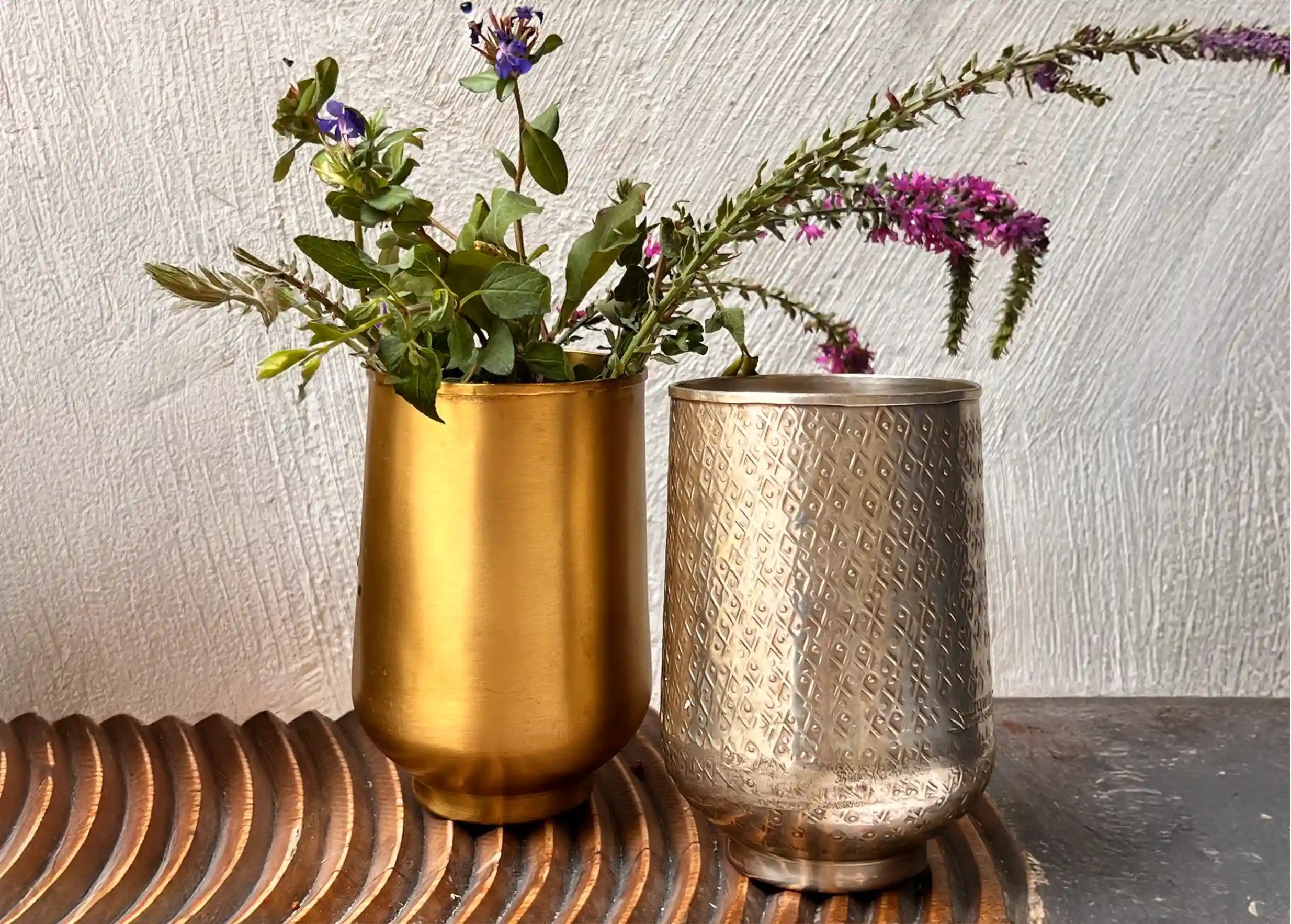 Two brass glasses on a wooden surface, one in gilded brass containing a bouquet of flowers, the other in silvered brass.