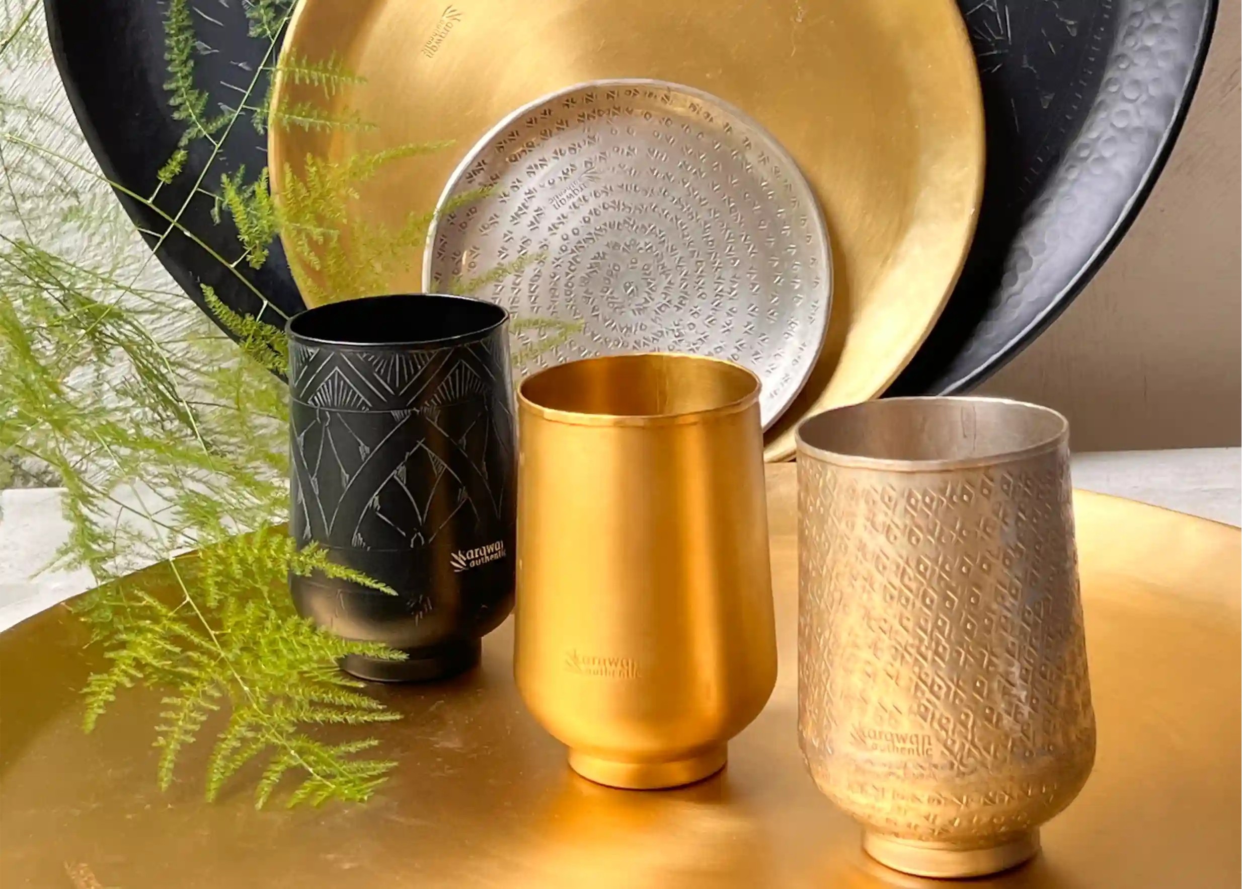 Trio de verres gravés en laiton, un verre noir, un verre doré, un verre argenté, avec comme un fond la collection de plateaux