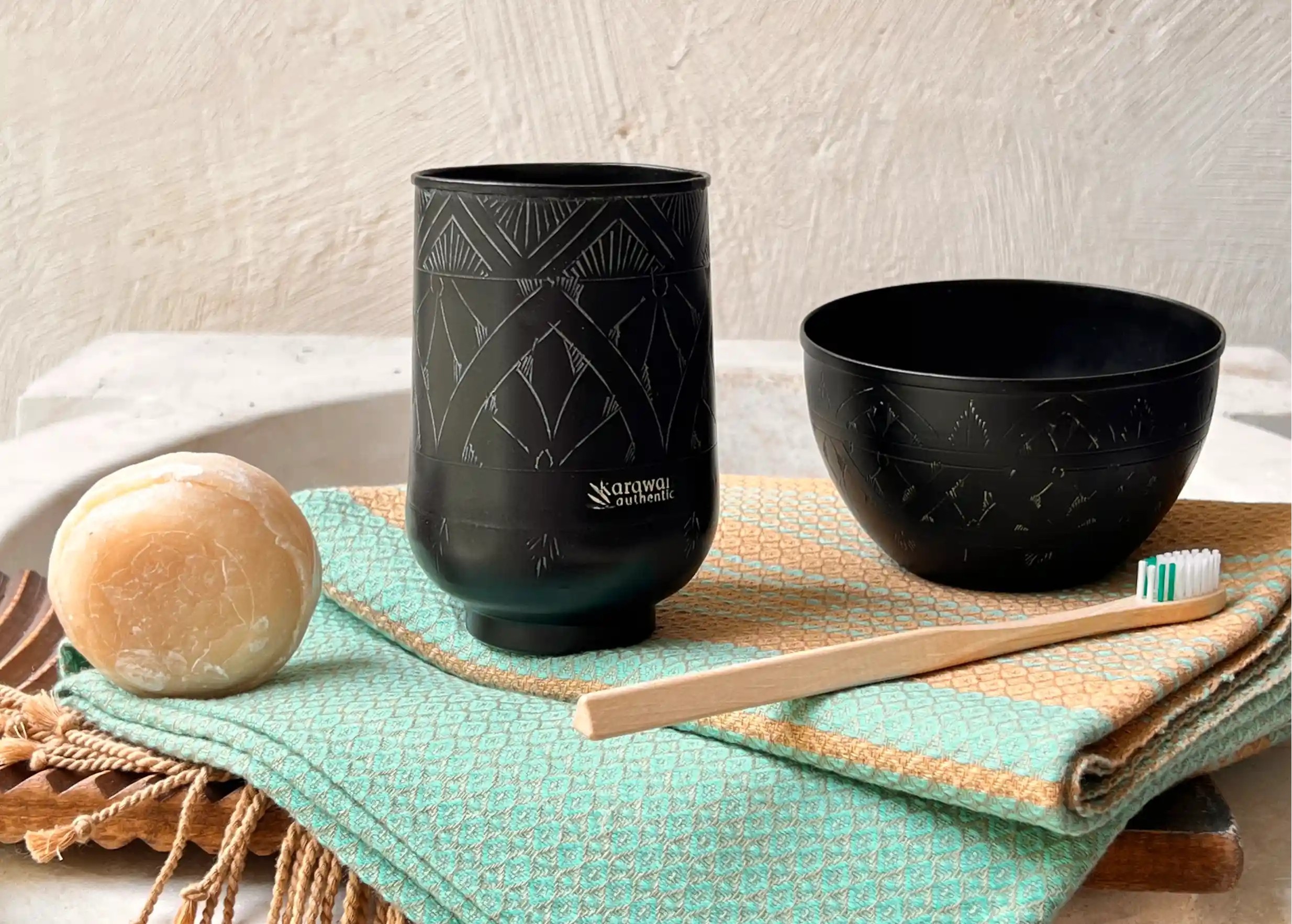 Verre en laiton noir avec des motifs gravés posé sur une fouta en coton bio, accompagné d'un savon rond et d'une brosse à dents