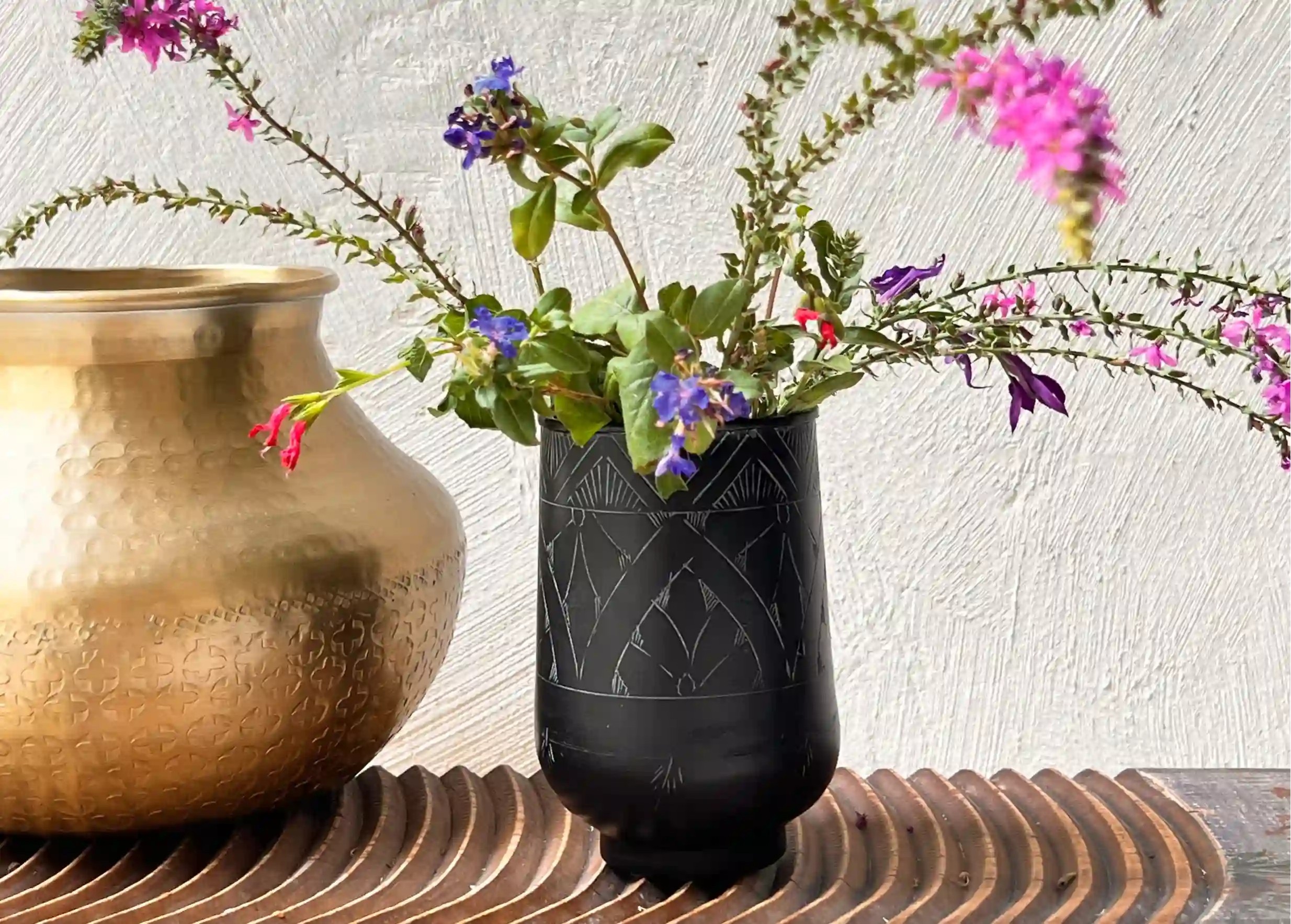 Verre noir gravé en laiton avec des motifs incrustés, un bouquet de fleurs colorées et des feuillages verts, posé sur du bois