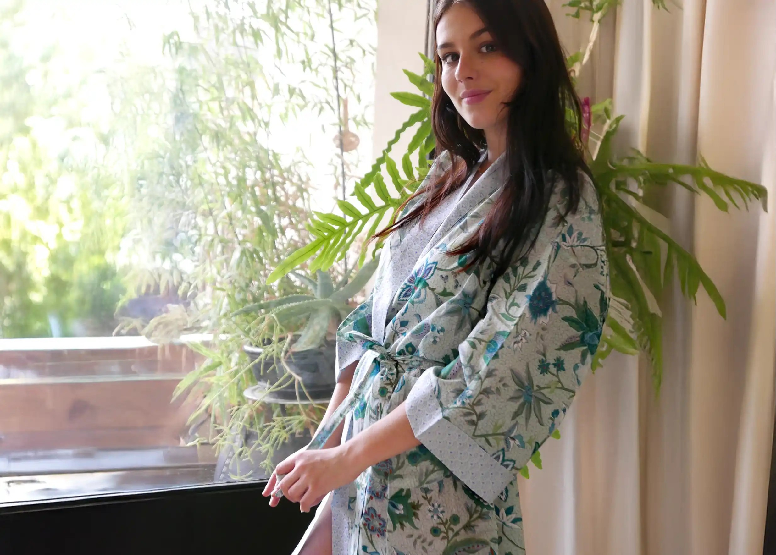 Woman wearing a Blue Garden cotton kimono in shades of blue and green, near a window with plants