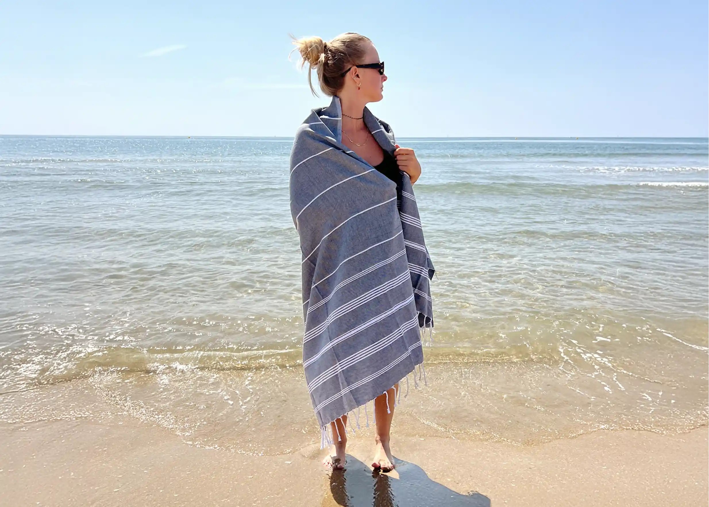 Femme portant une fouta anthracite en coton bio de la collection Hammam, devant une plage Femme portant une fouta anthracite en coton bio de la collection Hammam, devant une plage