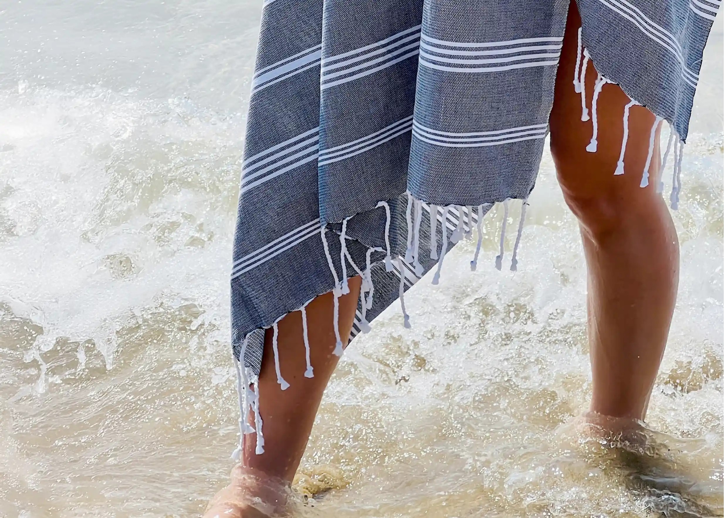 Femme portant une fouta anthracite en coton bio de la collection Hammam, les pieds dans l'eau Femme portant une fouta anthracite en coton bio de la collection Hammam, les pieds dans l'eau