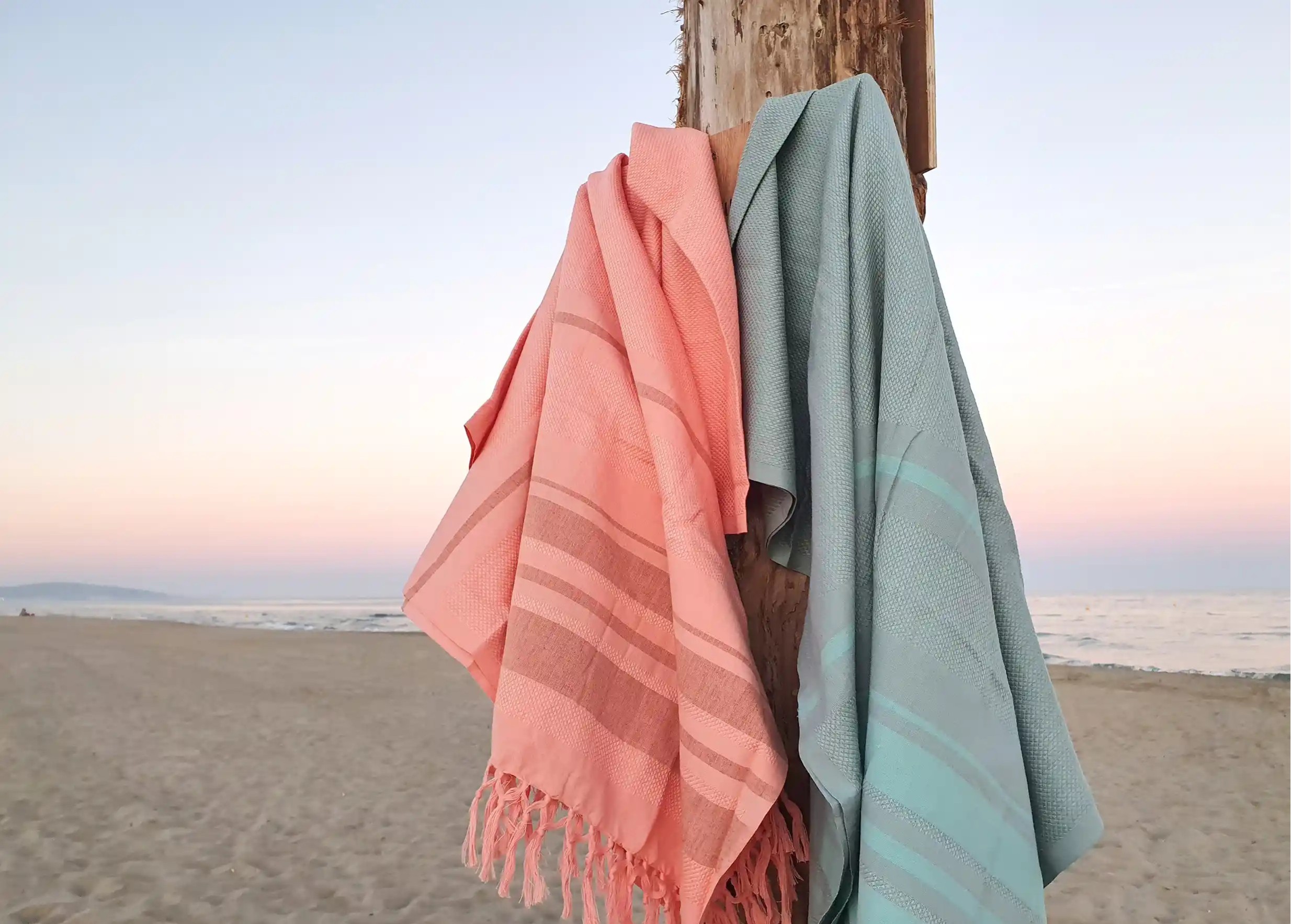 Two Garden foutas in organic cotton, with a honeycomb weave, hanging from a post on a beach, the sea in the distance.