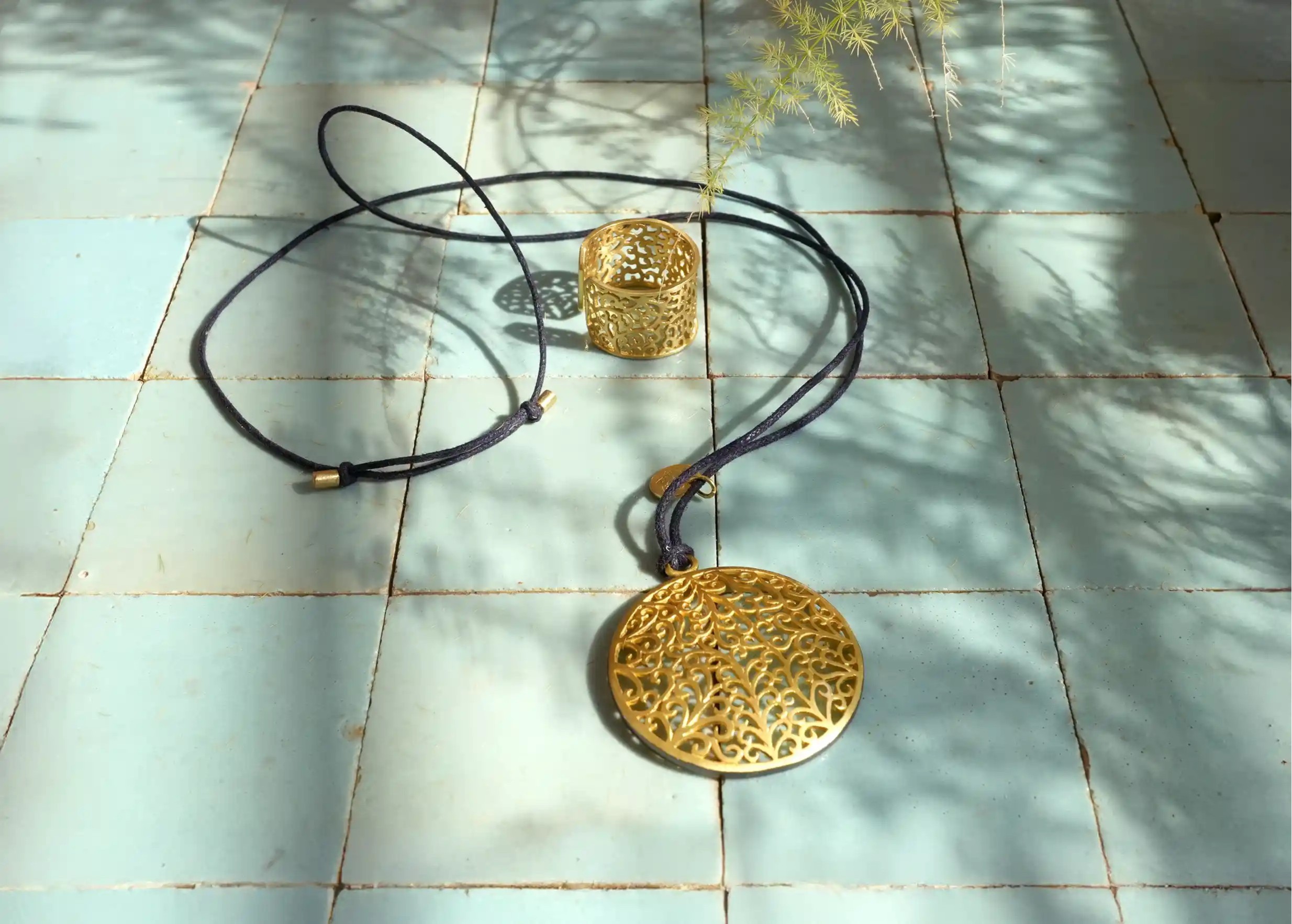 Pendentif en laiton de la gamme Jardin secret avec cordon noir, vus en hauteur sur une surface à carreaux