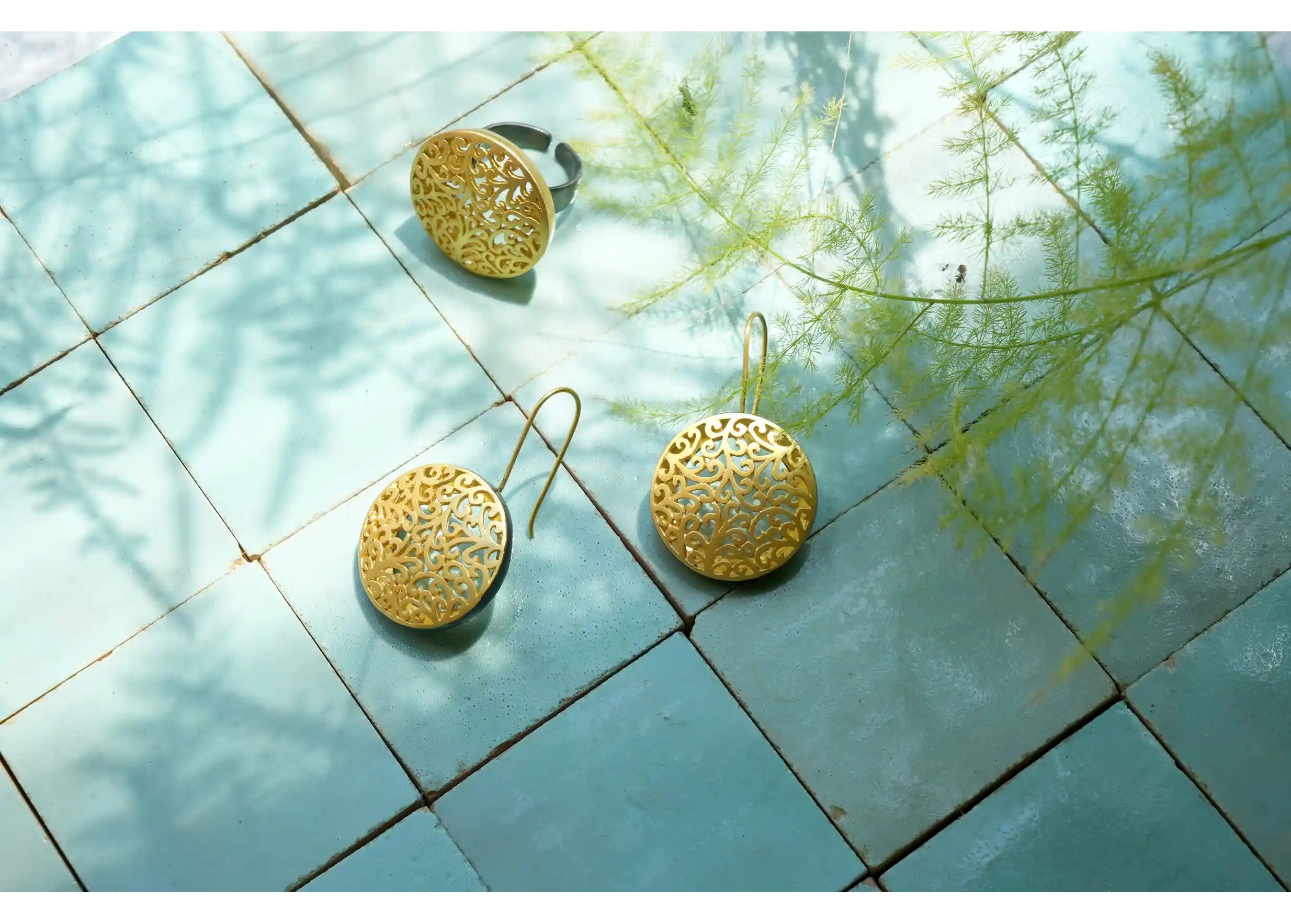 Boucles d'oreilles et bague en laiton de la gamme Jardin secret, posées sur des carreaux bleus